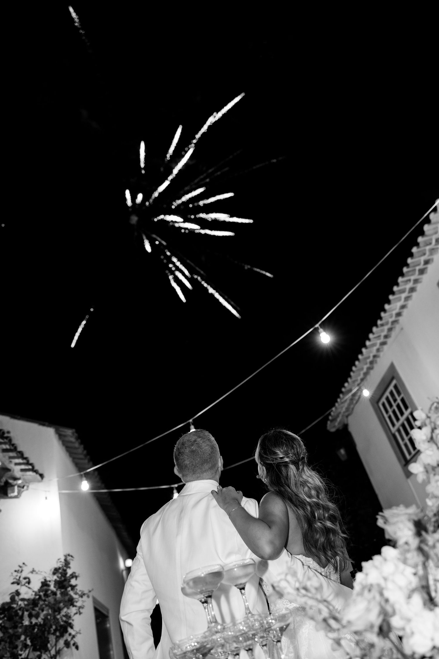Black and white photo of a couple watching fireworks at their outdoor wedding reception with string lights overhead.