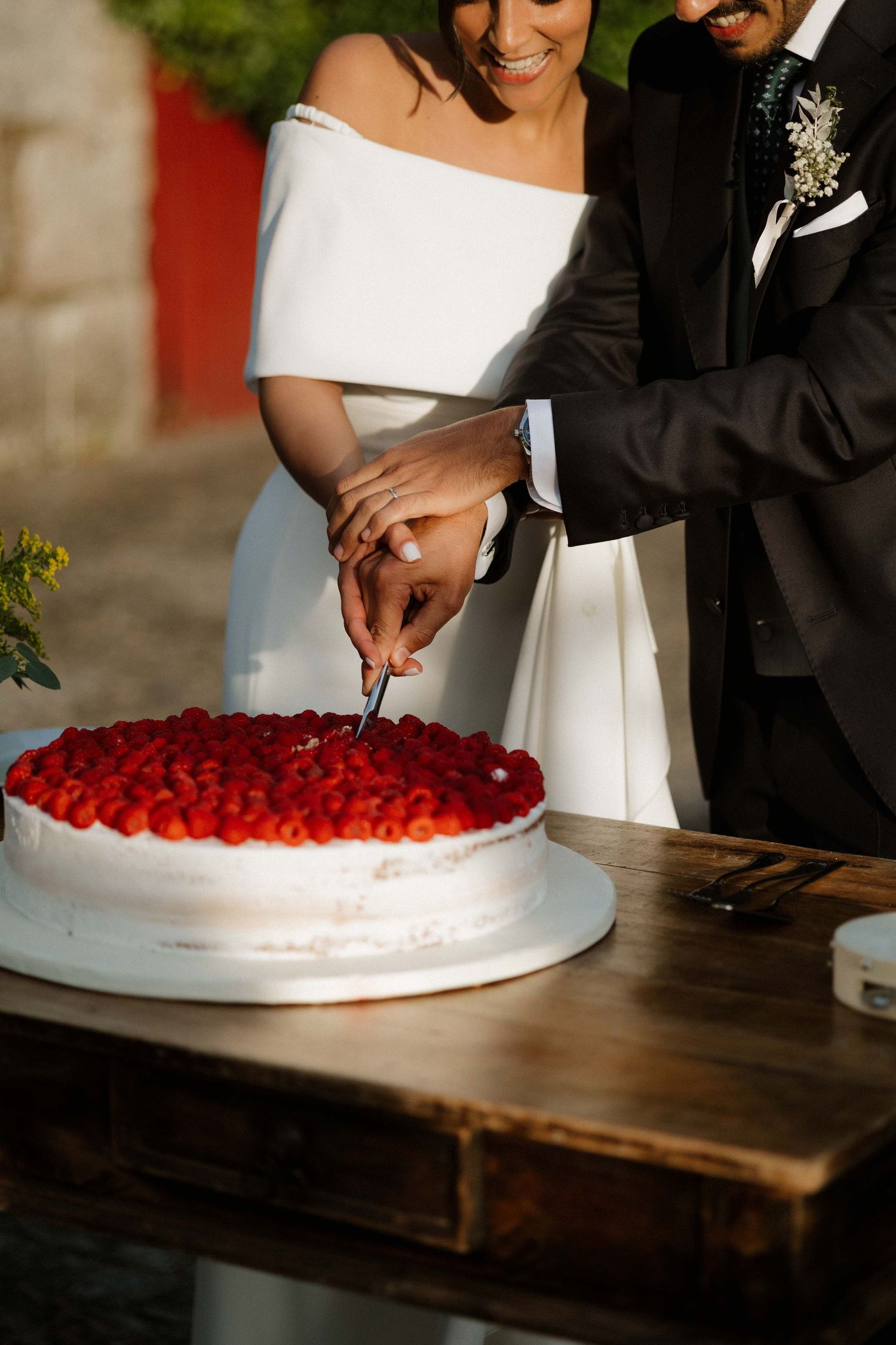 Bride and groom cutting a white wedding cake topped with fresh strawberries on a rustic wooden table outdoors.