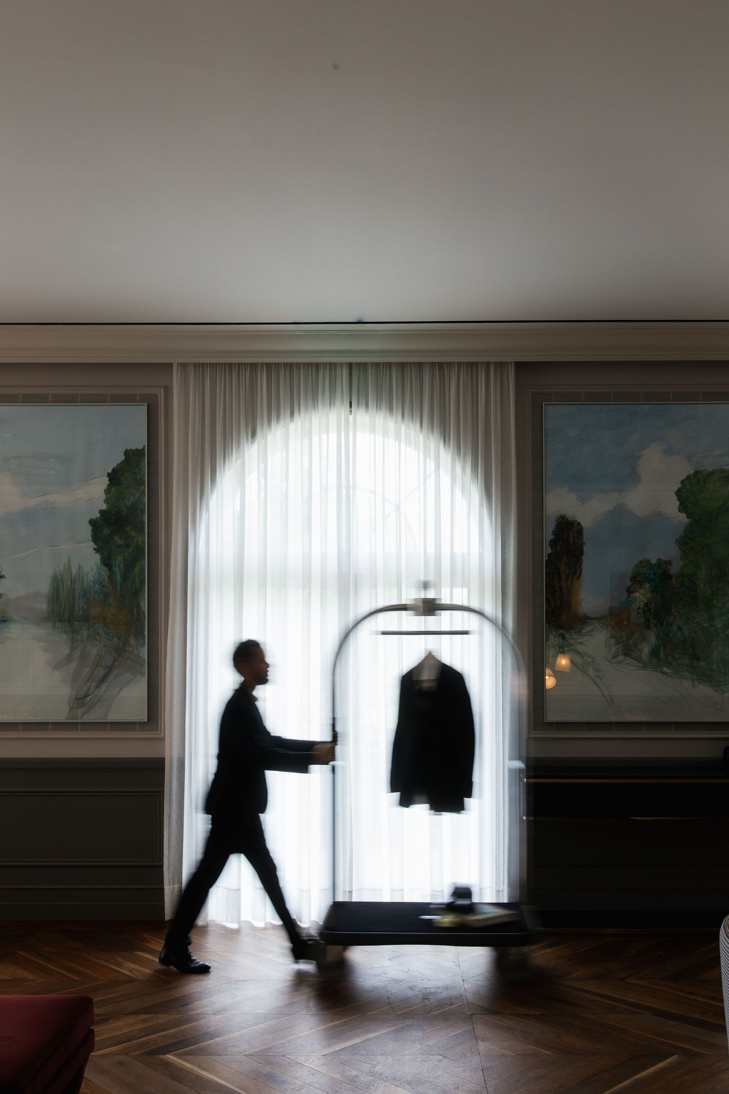 Silhouette of a hotel bellman pushing a luggage cart with a suit past an arched window.