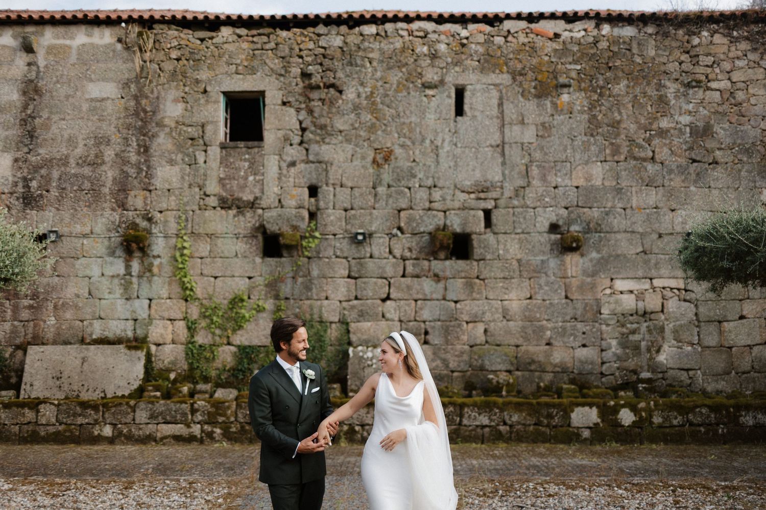 Newlyweds hold hands and smile at each other before rustic stone building during outdoor wedding portraits.