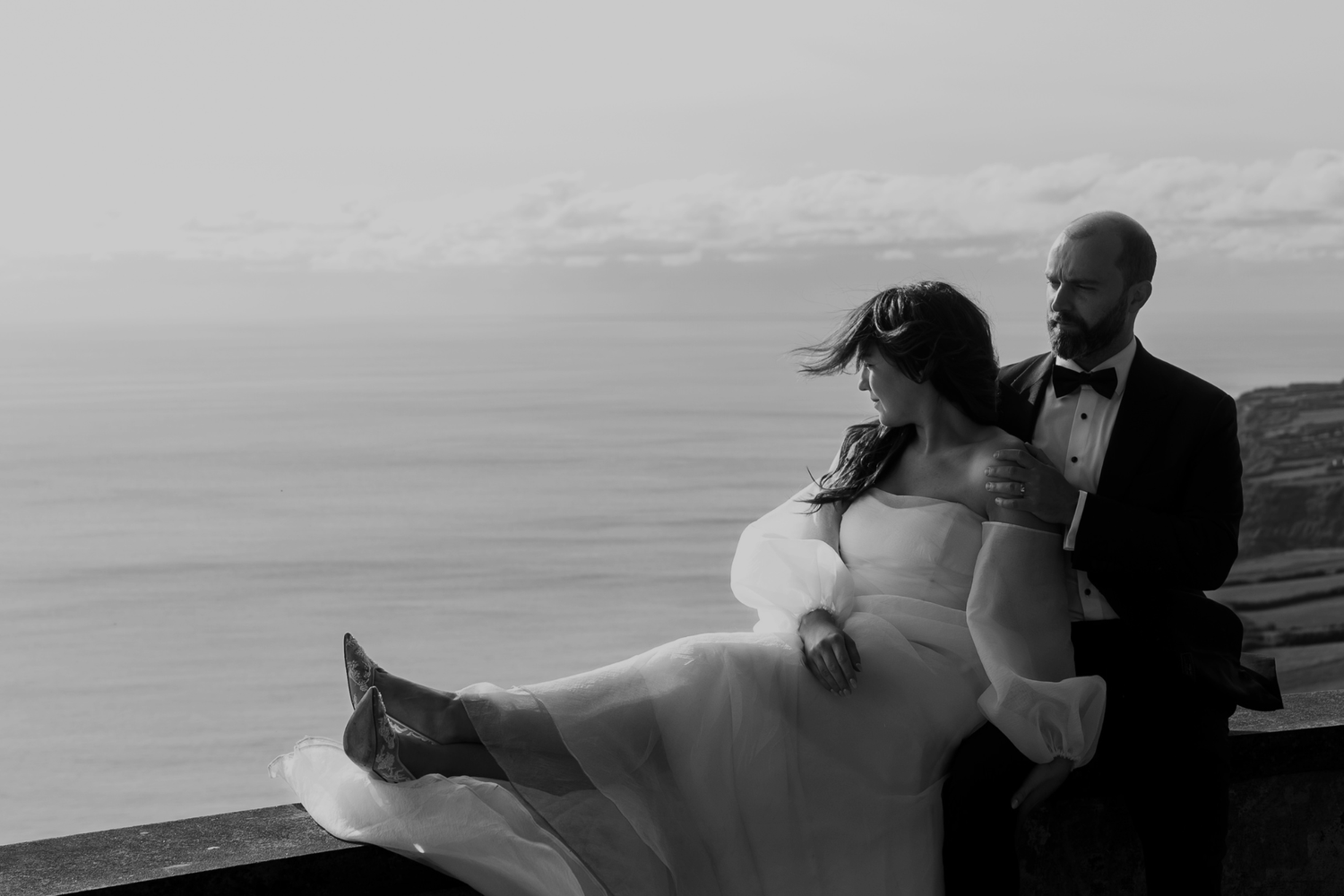 Black and white photo of a groom holding his bride on a cliffside overlooking the ocean.