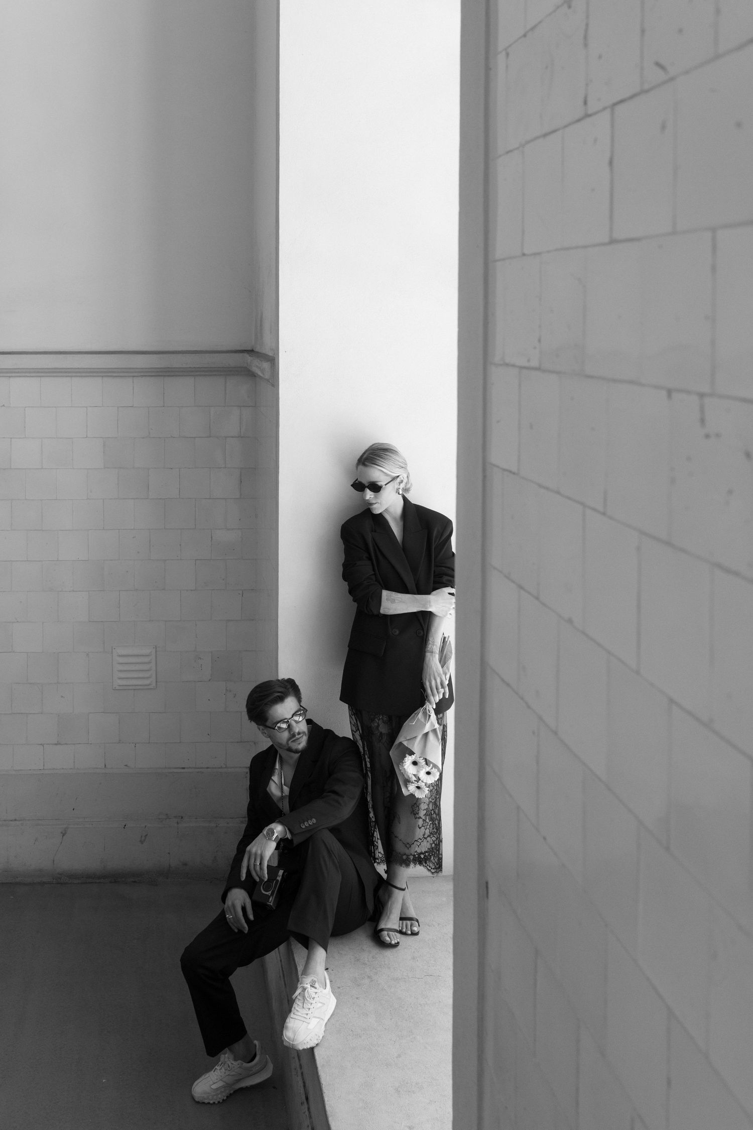Black and white portrait of a stylish couple in dark suits posing against a white brick wall outdoors.