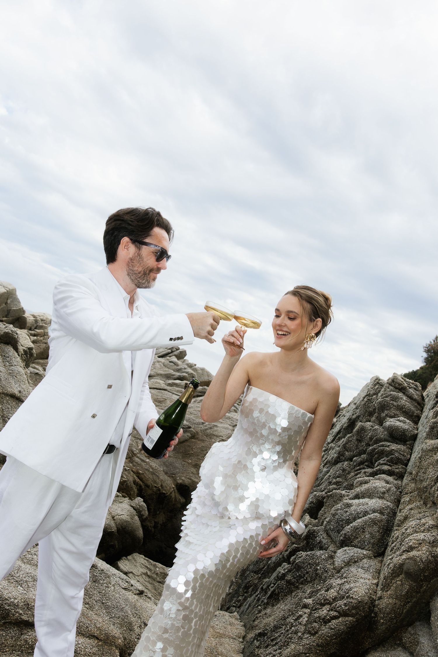 Groom in white suit toasting champagne with bride in sequin dress on dramatic rocky coastal backdrop.