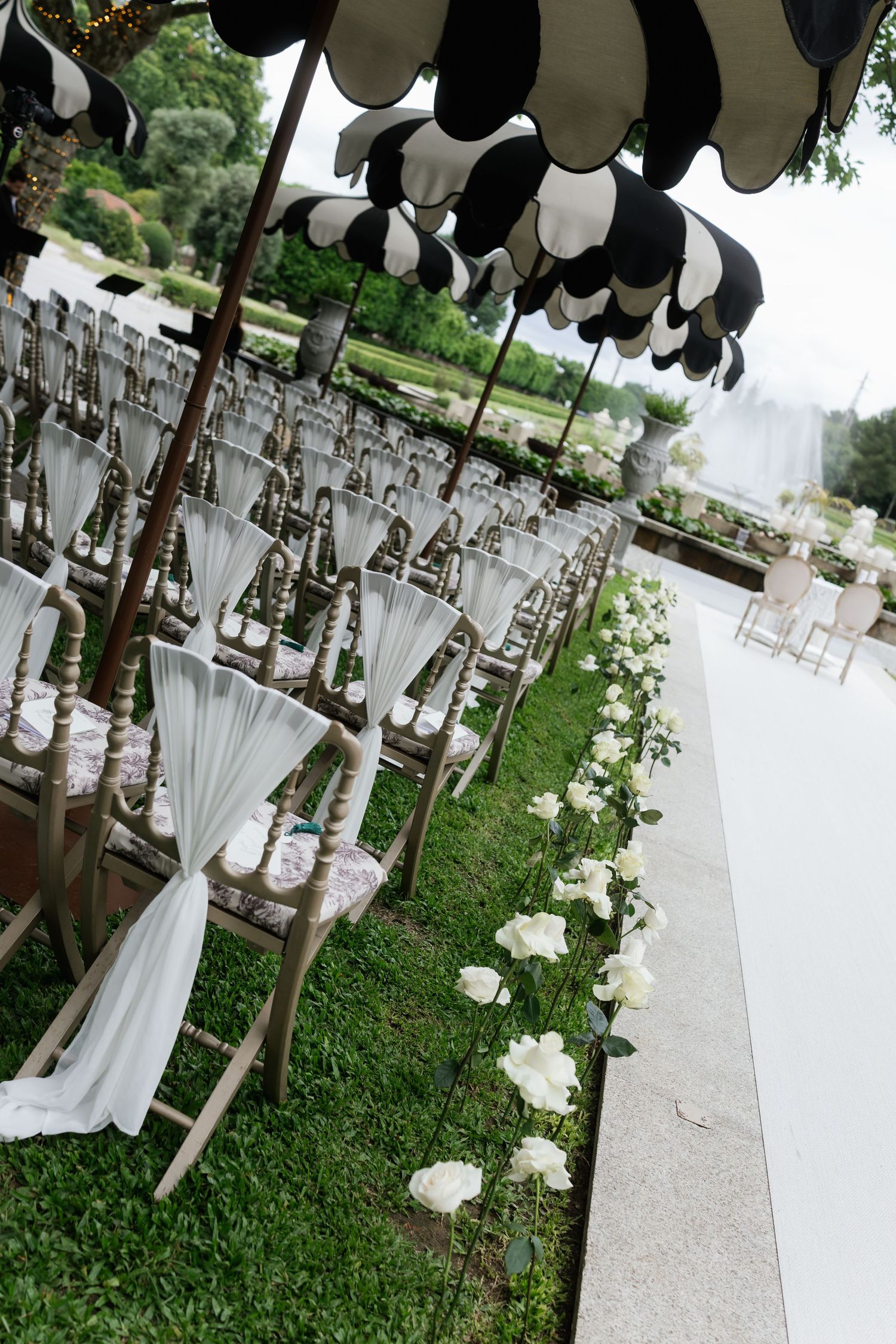 Outdoor wedding ceremony aisle lined with white roses and chiavari chairs under black and white striped umbrellas.