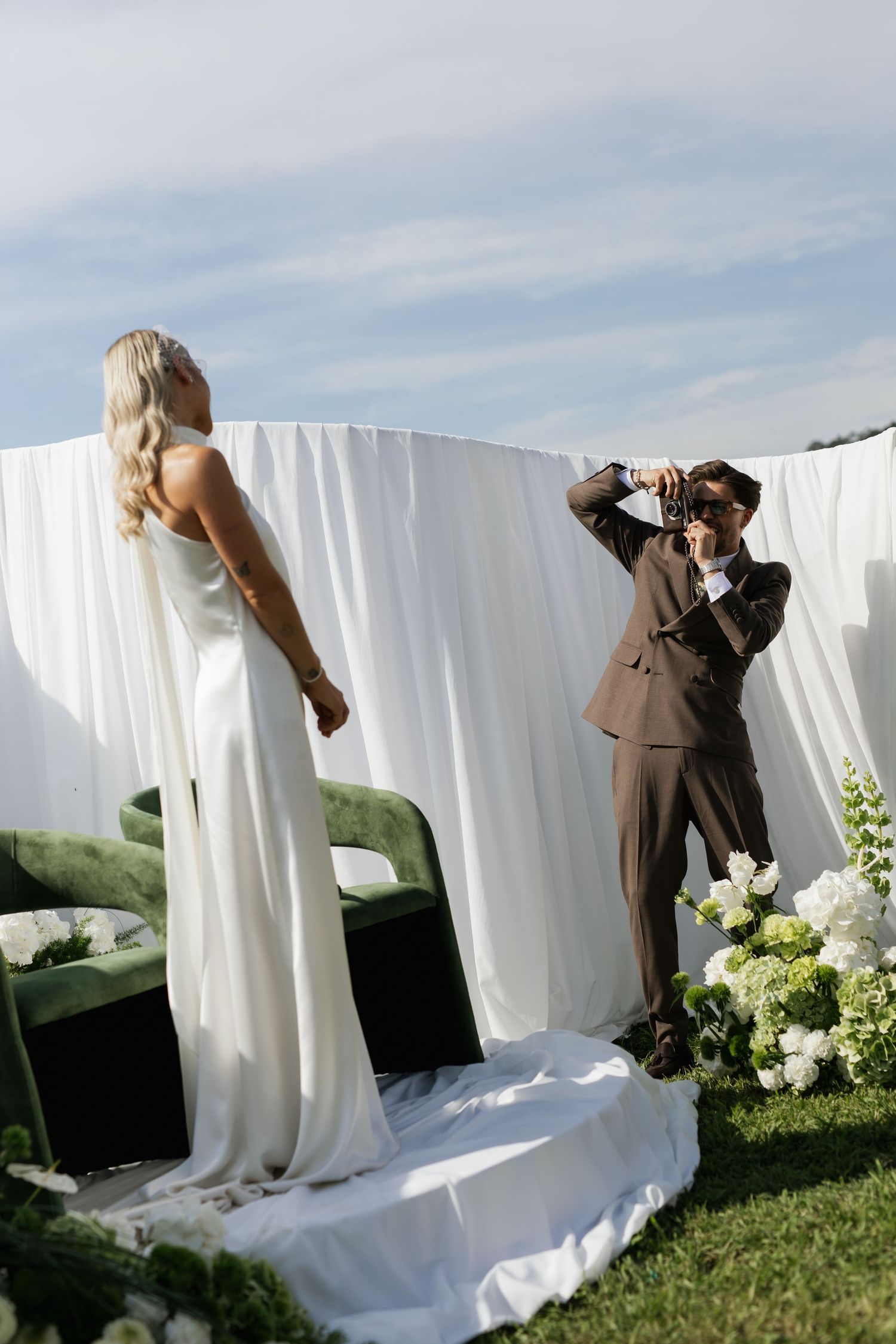 Wedding photographer in brown suit captures bride in white slip dress against white draped backdrop at outdoor ceremony.