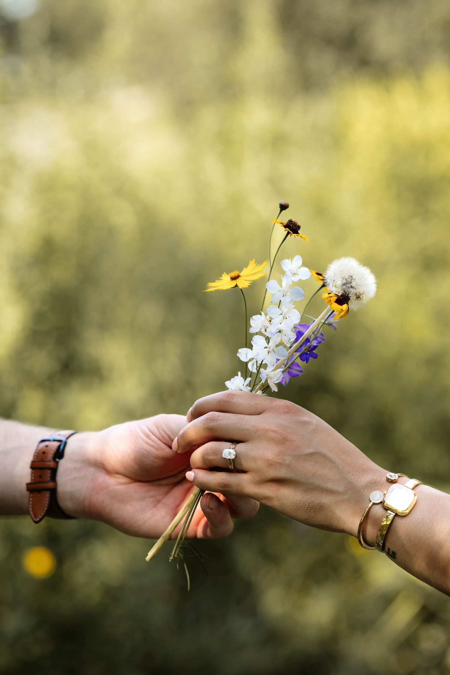 Two hands exchanging a small bouquet of wildflowers including yellow, white, and purple blooms with a dandelion outdoors.