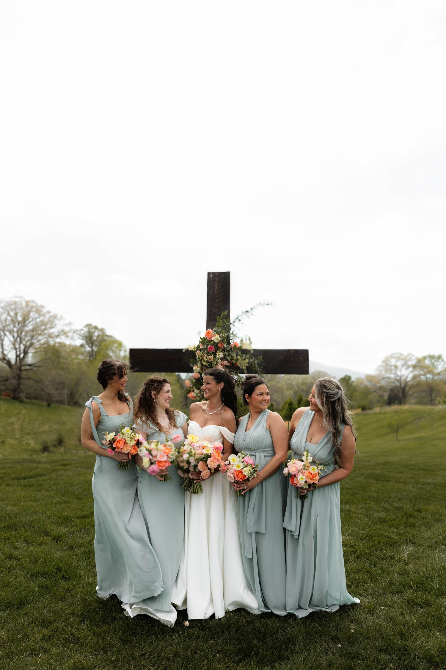 Bride in white gown with bridesmaids in sage green dresses holding colorful bouquets in front of a wooden cross outdoors.