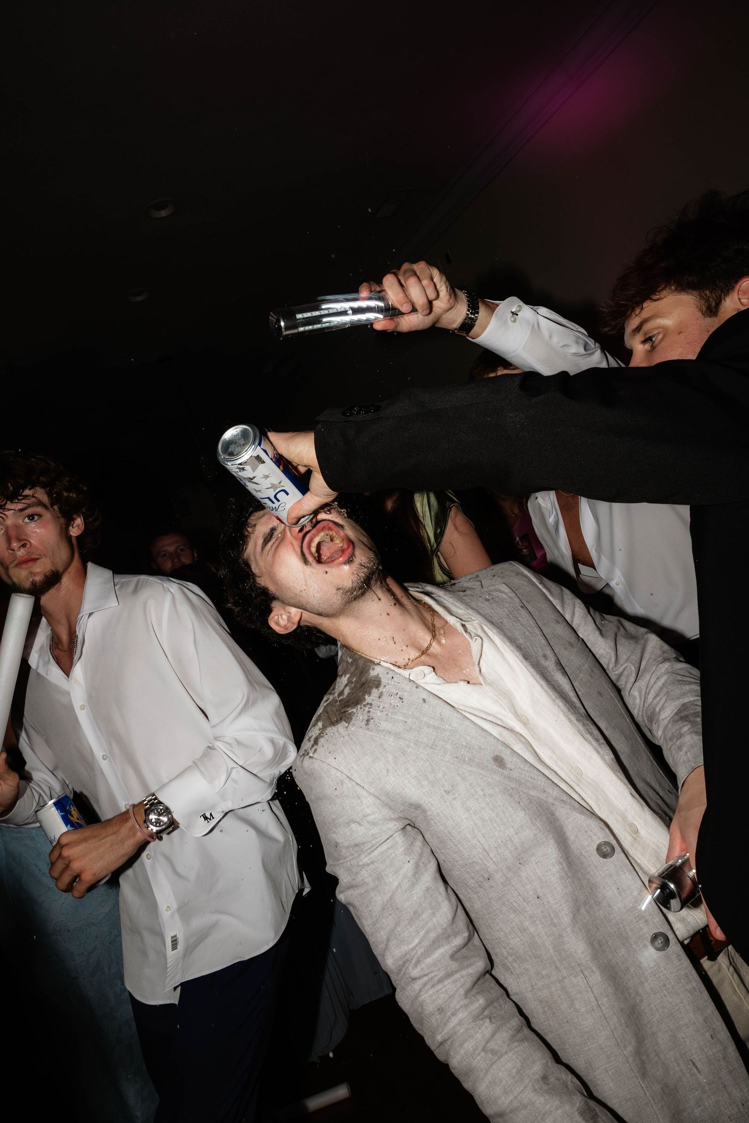 Young man tilts head back as friend pours drink into his mouth at a lively party.
