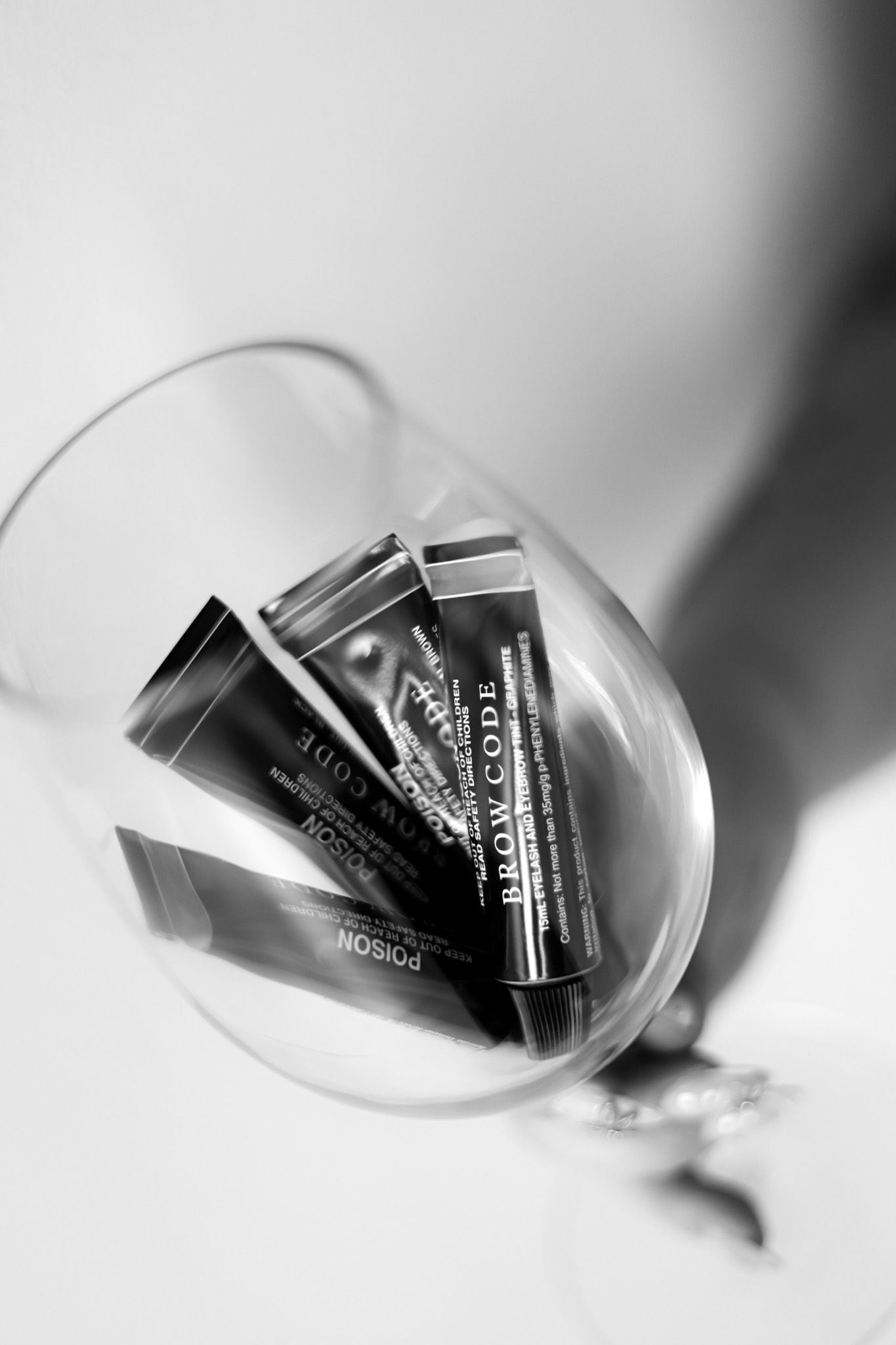 Black and white photo of Brow Code makeup tubes inside a tilted wine glass with dramatic shadows.