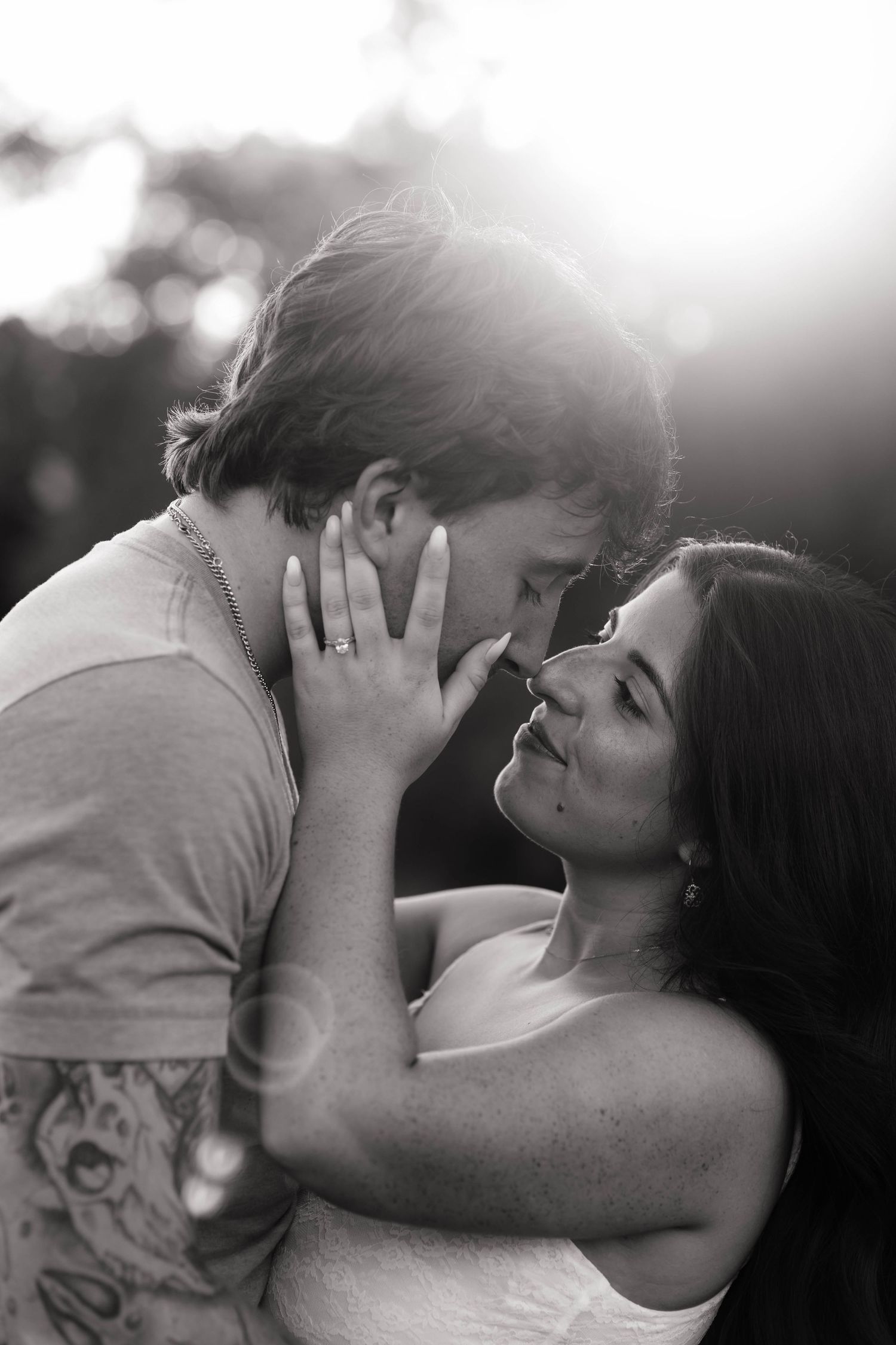 Black and white photo of a romantic couple almost kissing, woman wearing engagement ring, soft bokeh background.