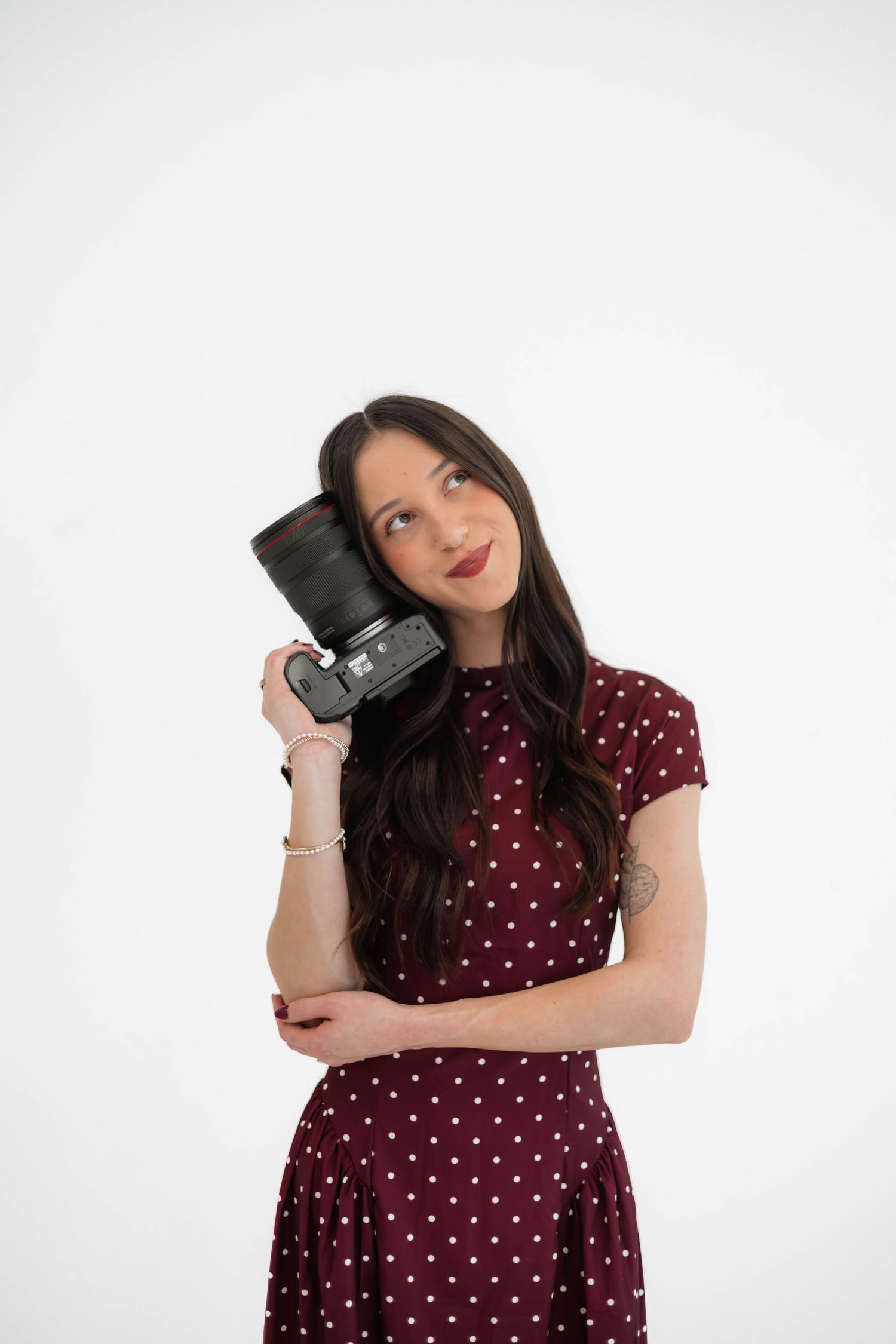 Young woman in burgundy polka dot dress holding a DSLR camera, posing against a white background.