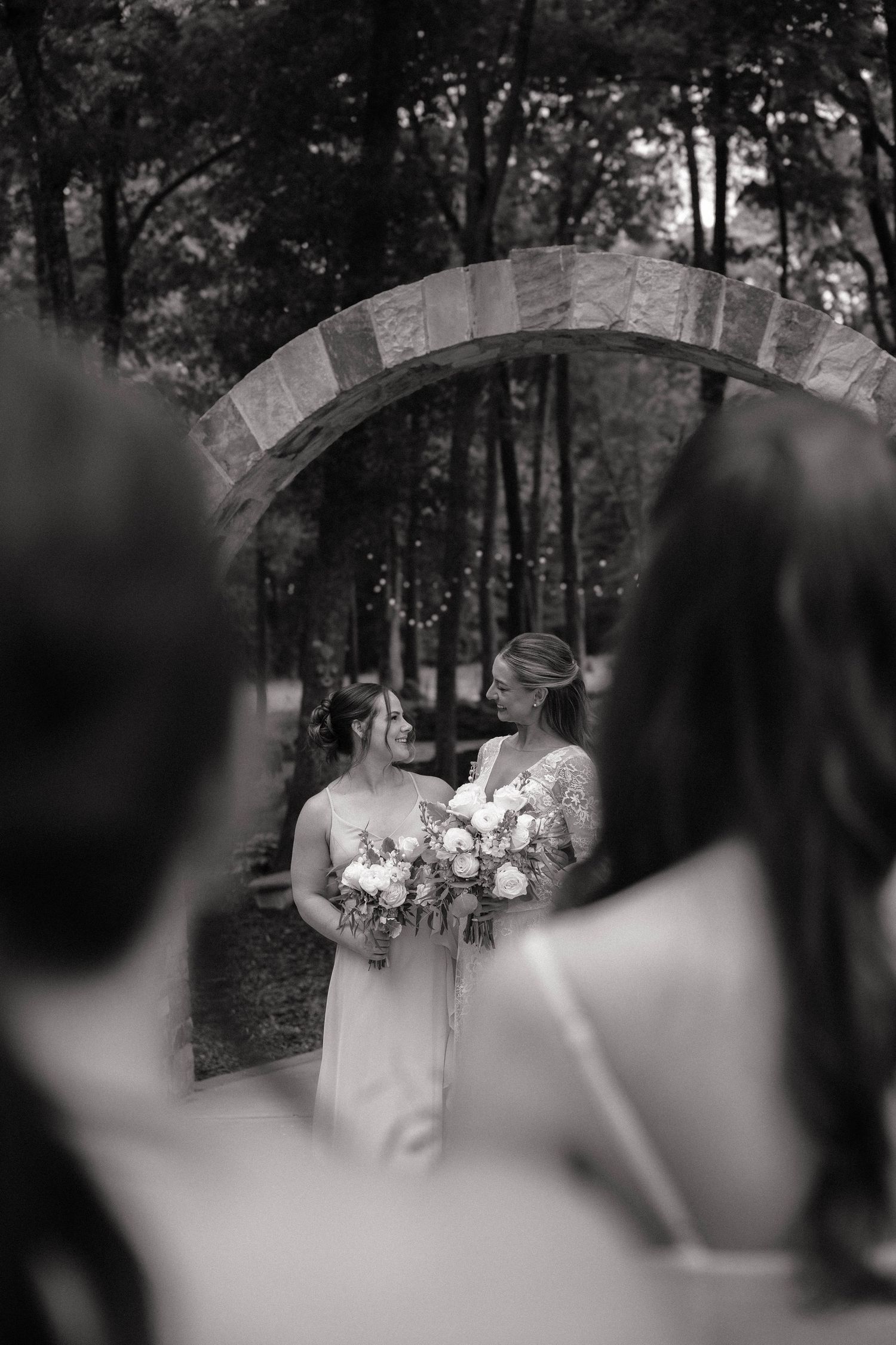 Black and white photo of two brides holding bouquets under a stone arch, exchanging glances at an outdoor wedding.