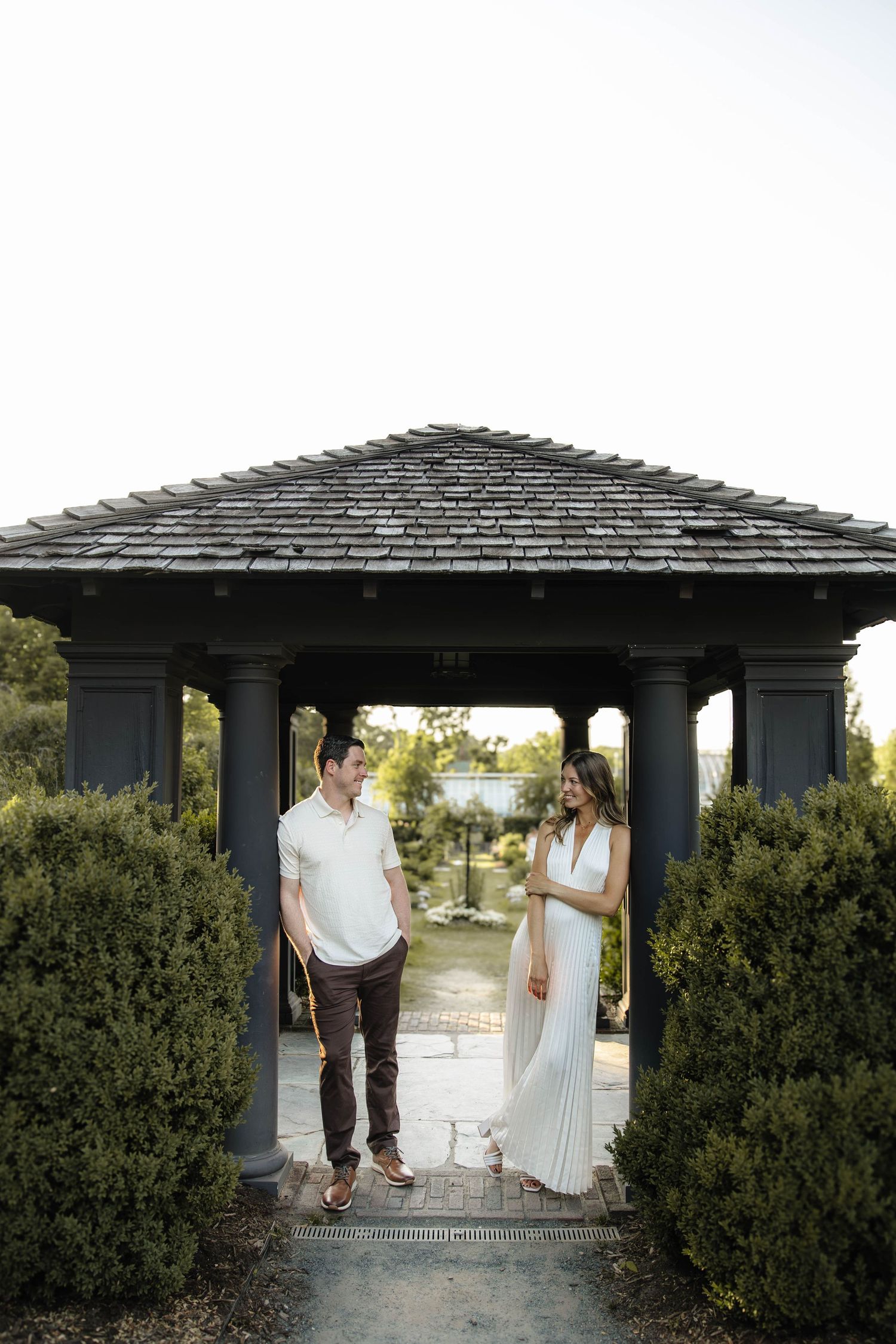 Couple standing under a dark gazebo surrounded by lush hedges in an outdoor garden setting.