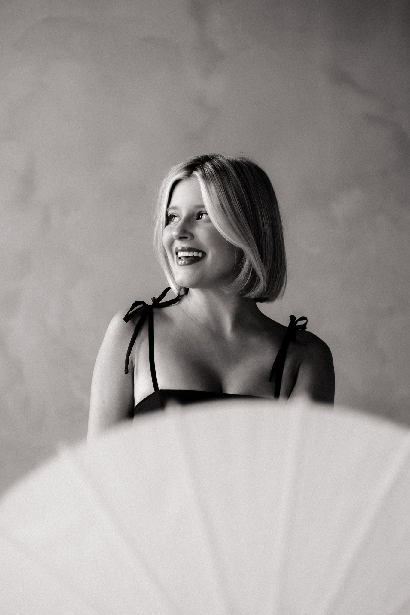 Black and white portrait of a smiling blonde woman in a strappy top holding a white fan against a marble background.