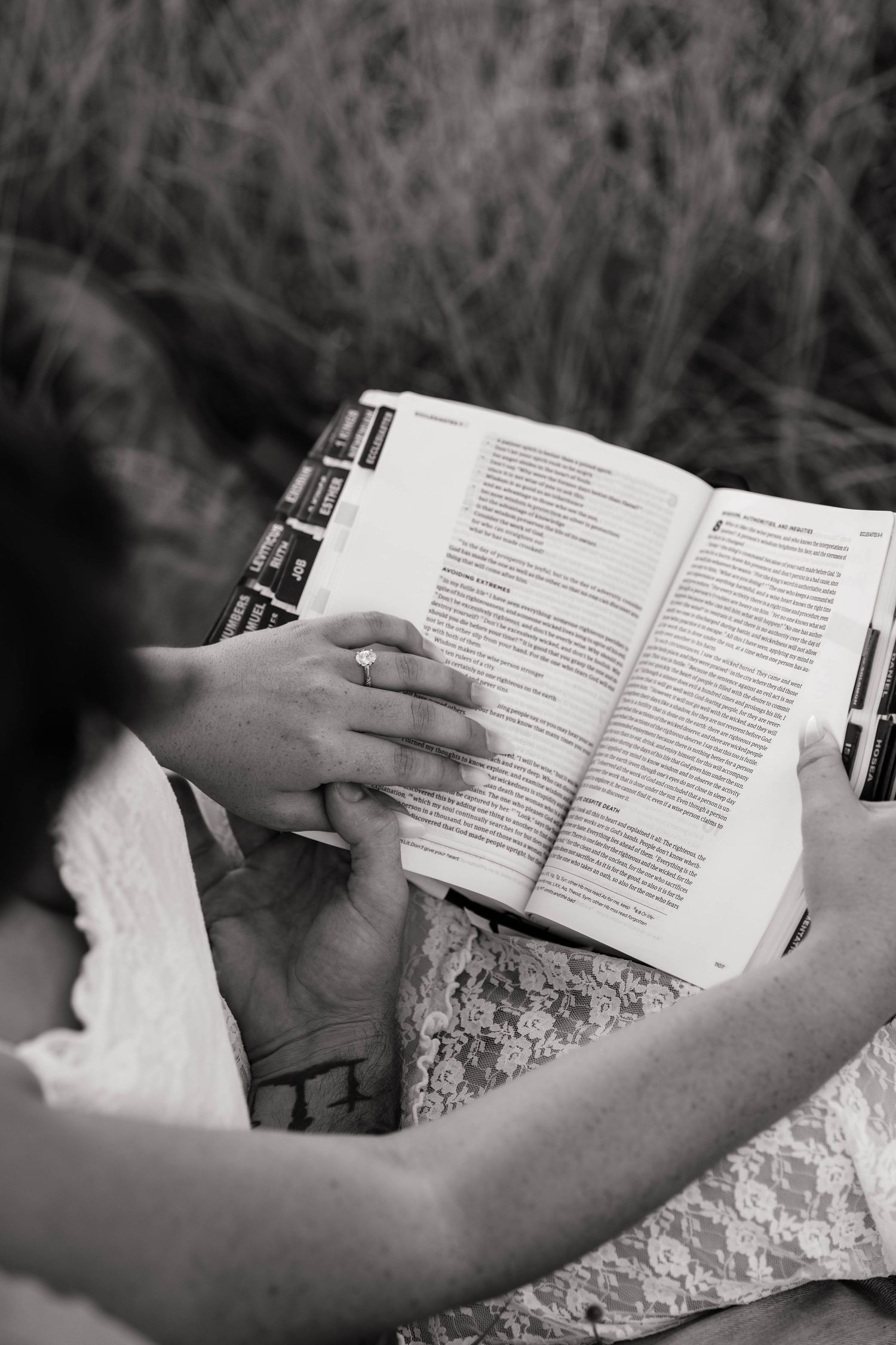 Black and white photo of couple holding hands over an open Bible outdoors.
