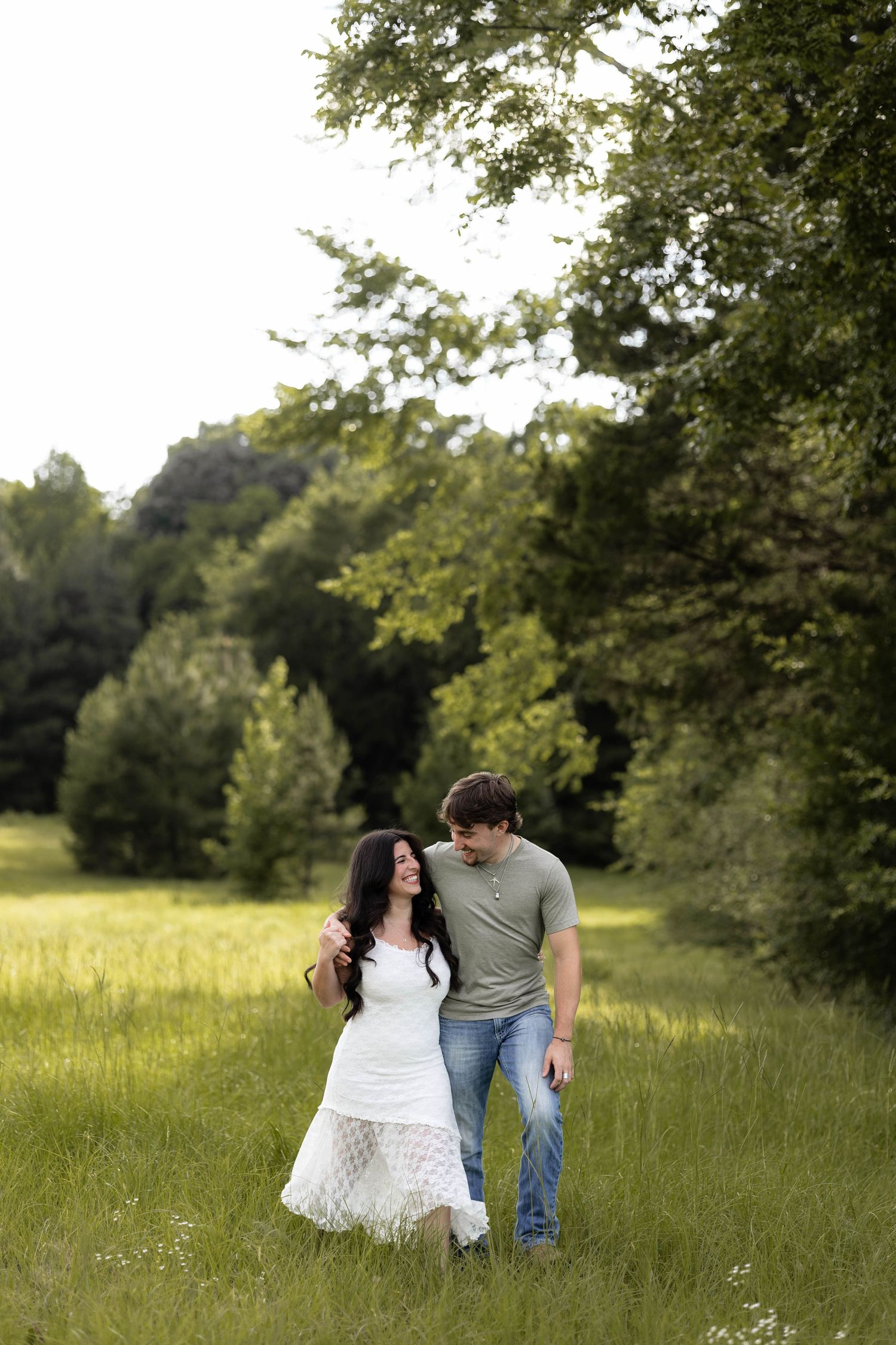 A couple walks hand-in-hand through a lush green meadow, the woman in a white dress and man in a gray shirt and jeans.