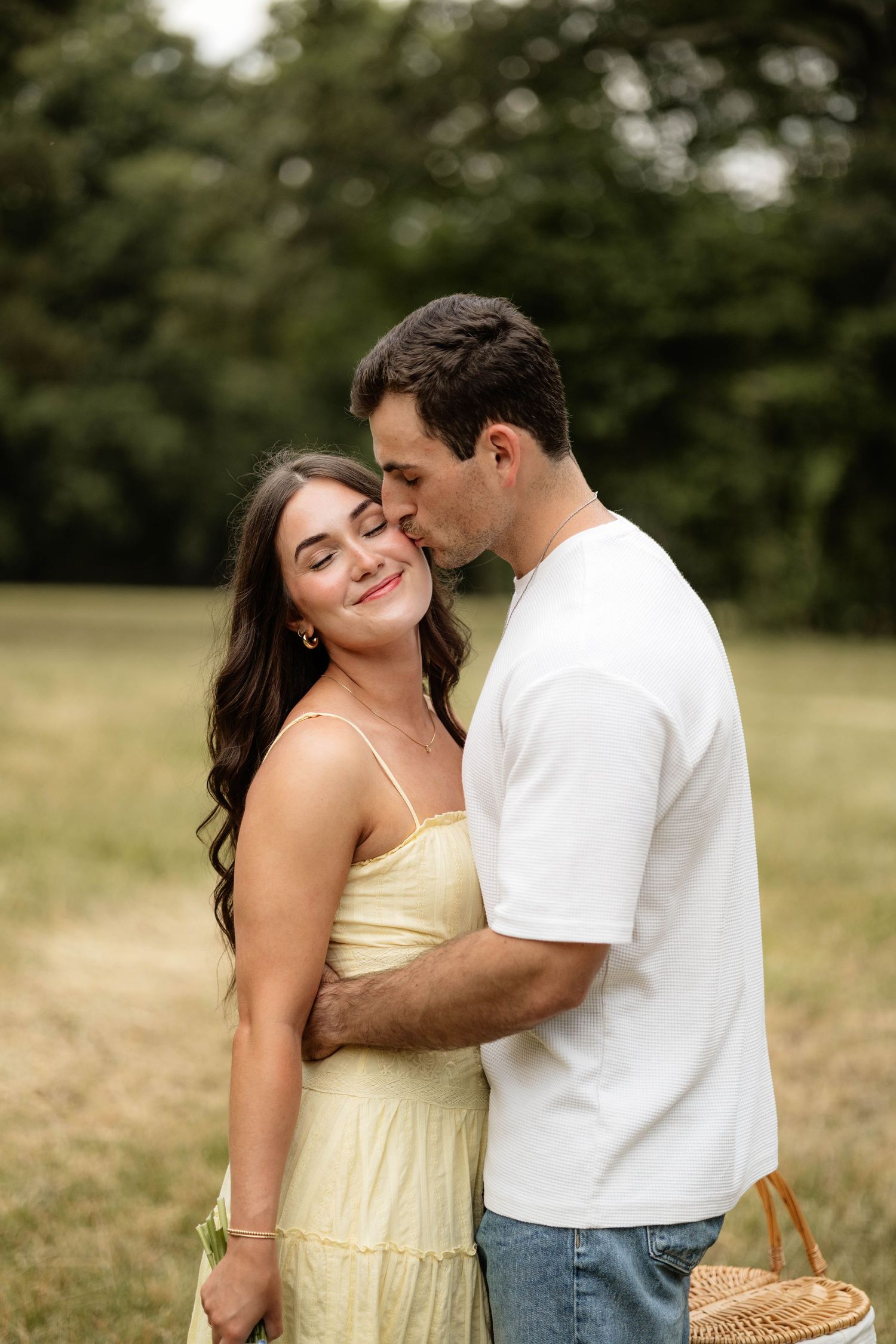 A couple shares a tender moment outdoors, man kissing woman's cheek as she smiles in a yellow dress.