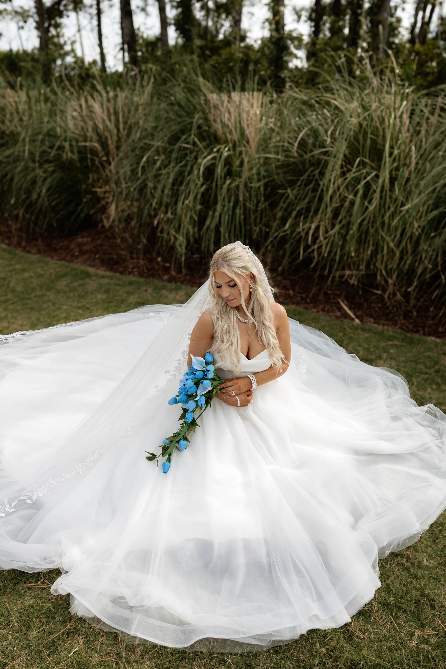 Blonde bride twirls in flowing white ballgown holding blue floral bouquet in outdoor garden setting.