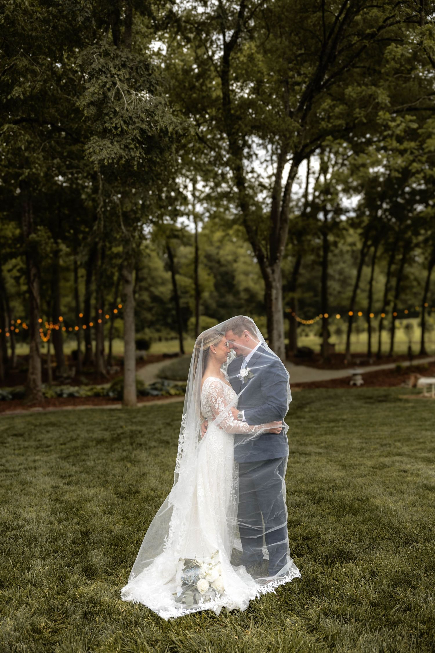 Bride and groom sharing a romantic moment under a flowing veil on a lush green lawn surrounded by trees.