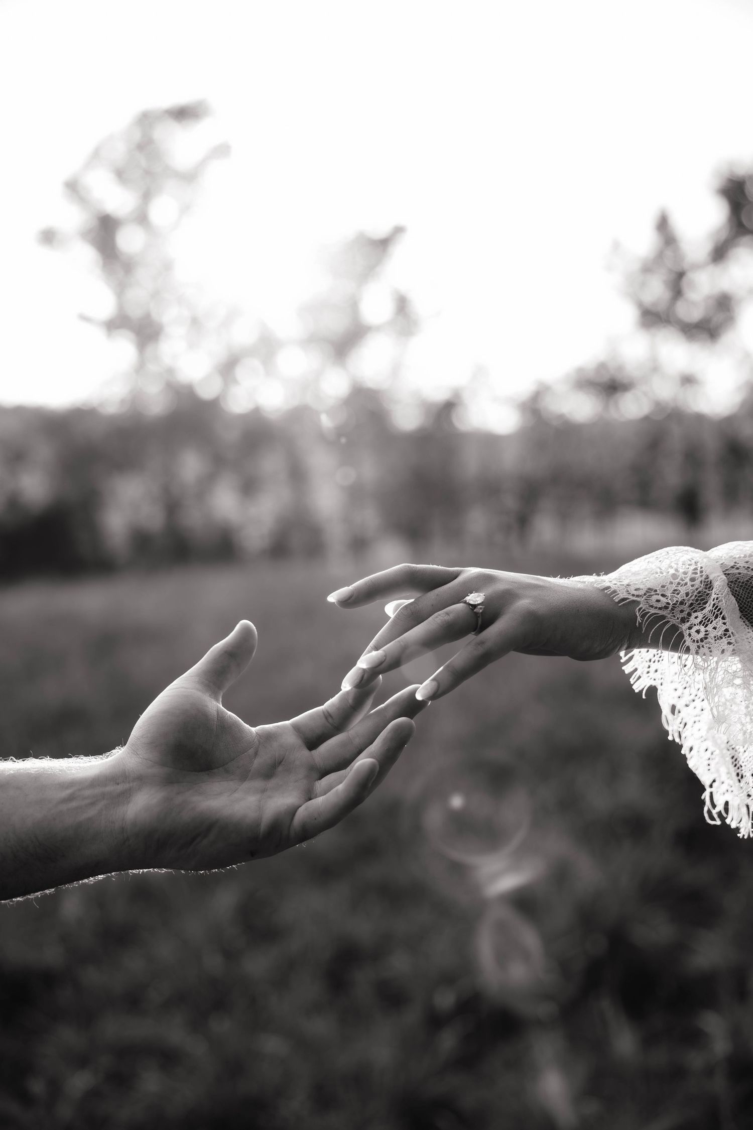 Black and white photo of a couple's hands almost touching, with a wedding ring visible, set against a blurred outdoor background.
