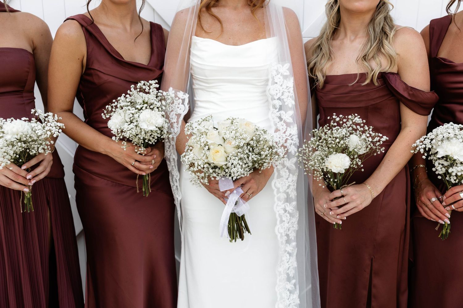 Bride in white gown with bridesmaids in burgundy satin dresses holding white floral bouquets with baby's breath.