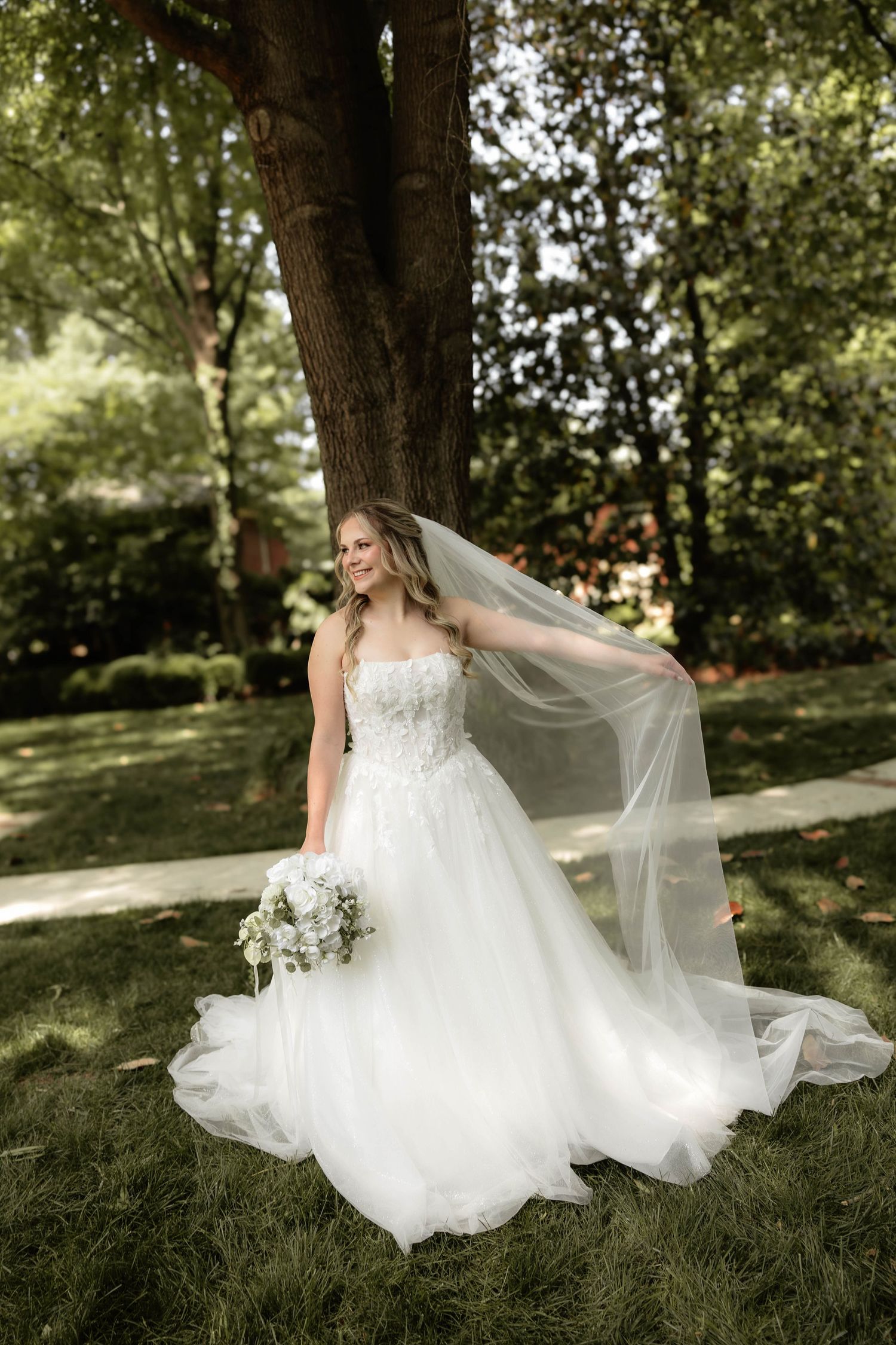Bride in white lace ballgown with flowing veil holds bouquet outdoors under trees on green grass.