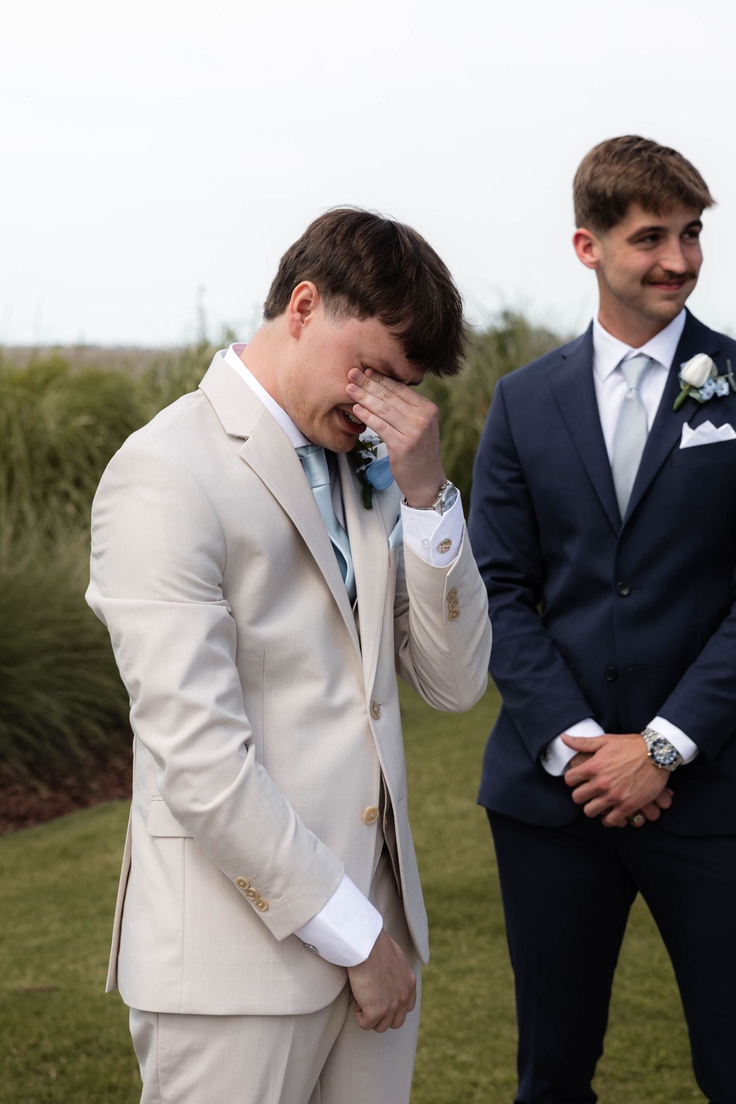 Emotional groom in beige suit crying during wedding ceremony while best man in navy suit stands beside him.