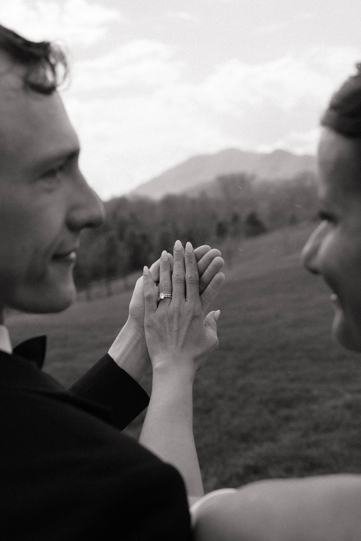 Black and white photo of smiling bride and groom holding hands, showing wedding ring, with mountain landscape background.