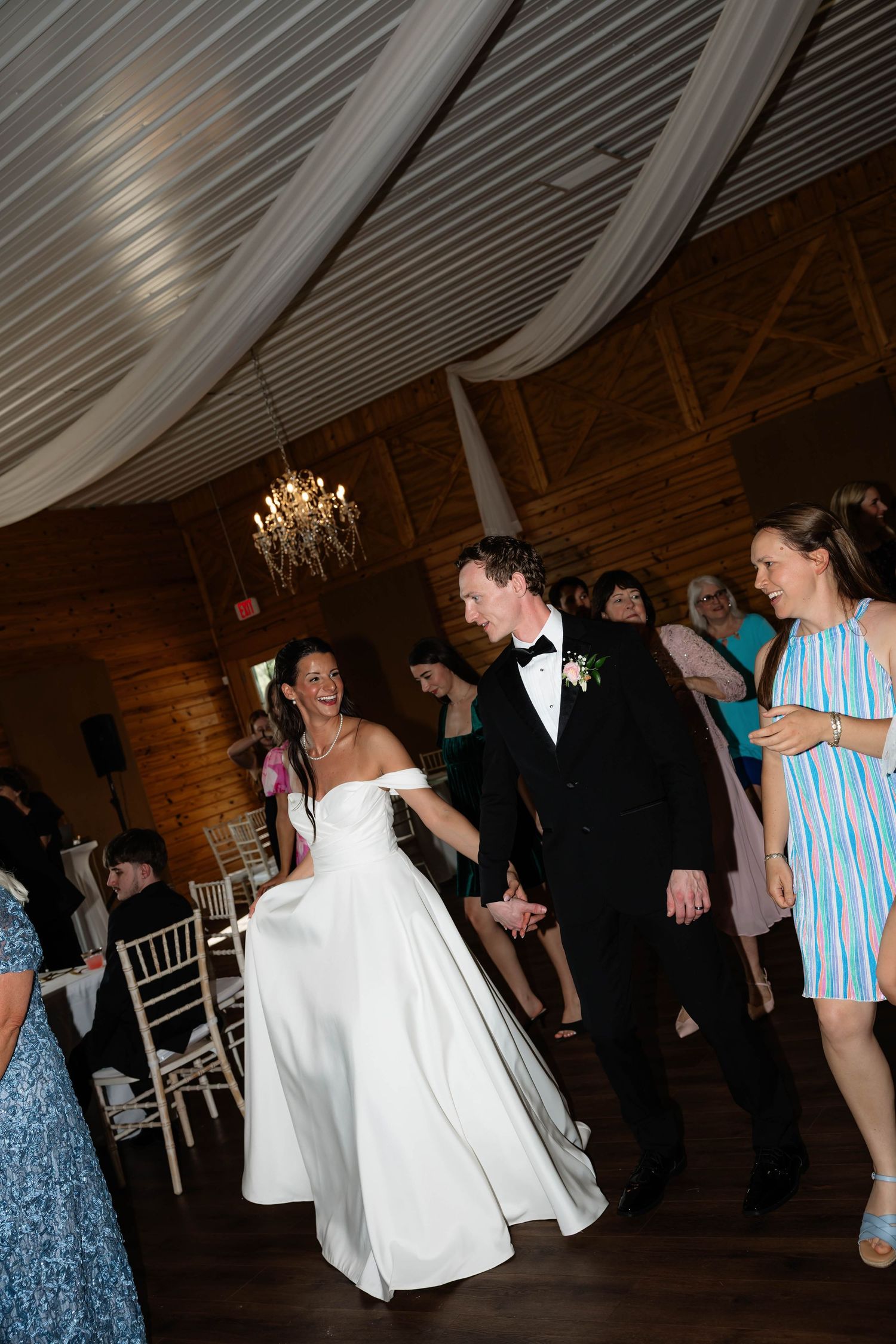 Bride in white off-shoulder gown and groom in black tuxedo share first dance at rustic barn wedding reception.