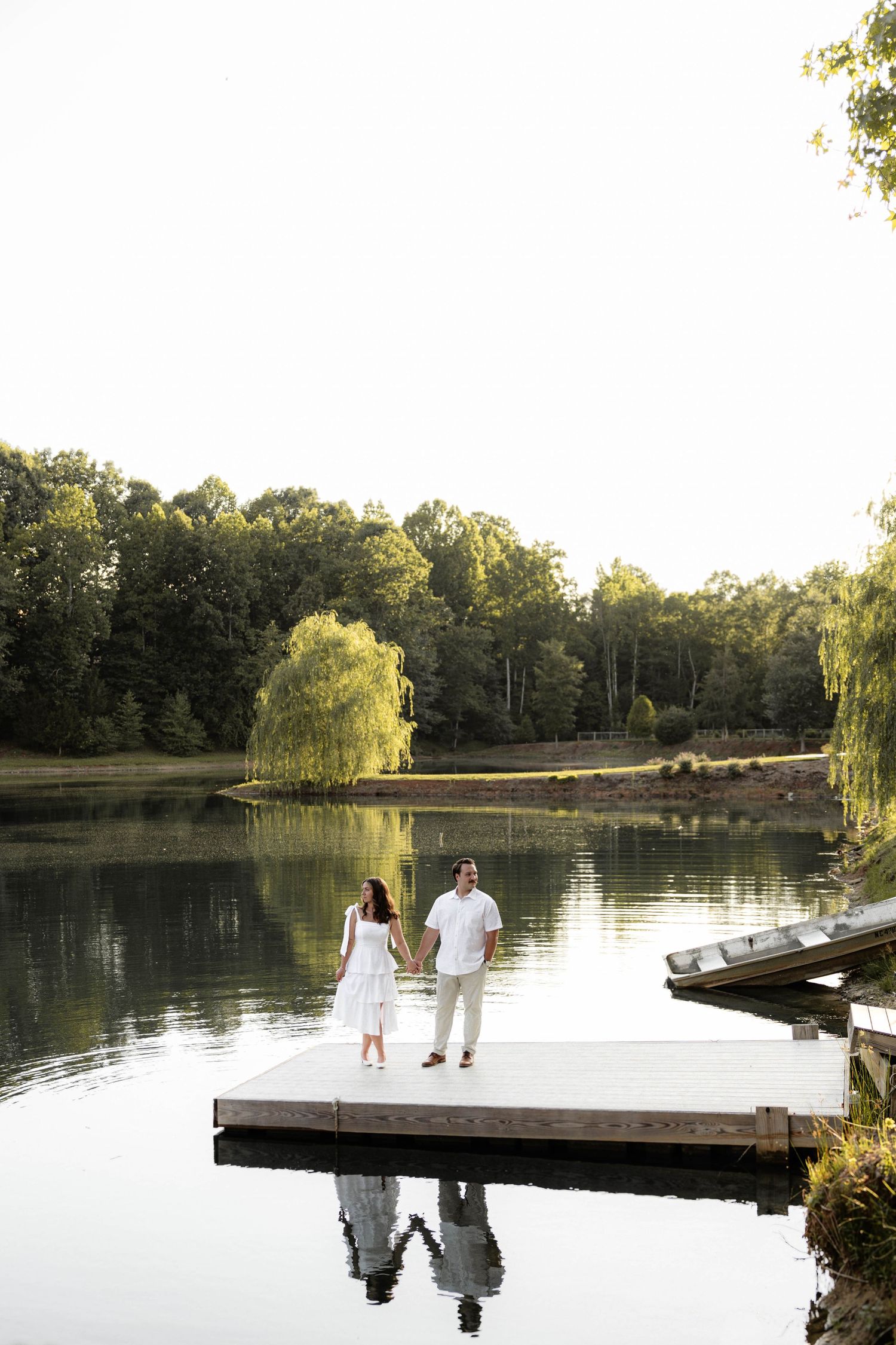 Couple in white holding hands on a lakeside dock surrounded by lush green trees and calm reflective water.
