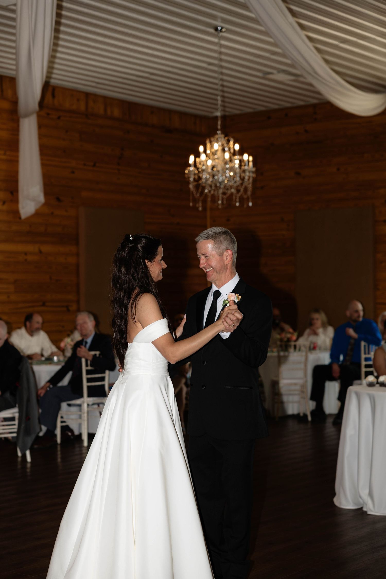 Bride and groom share their first dance under a chandelier in a rustic wooden wedding reception venue.