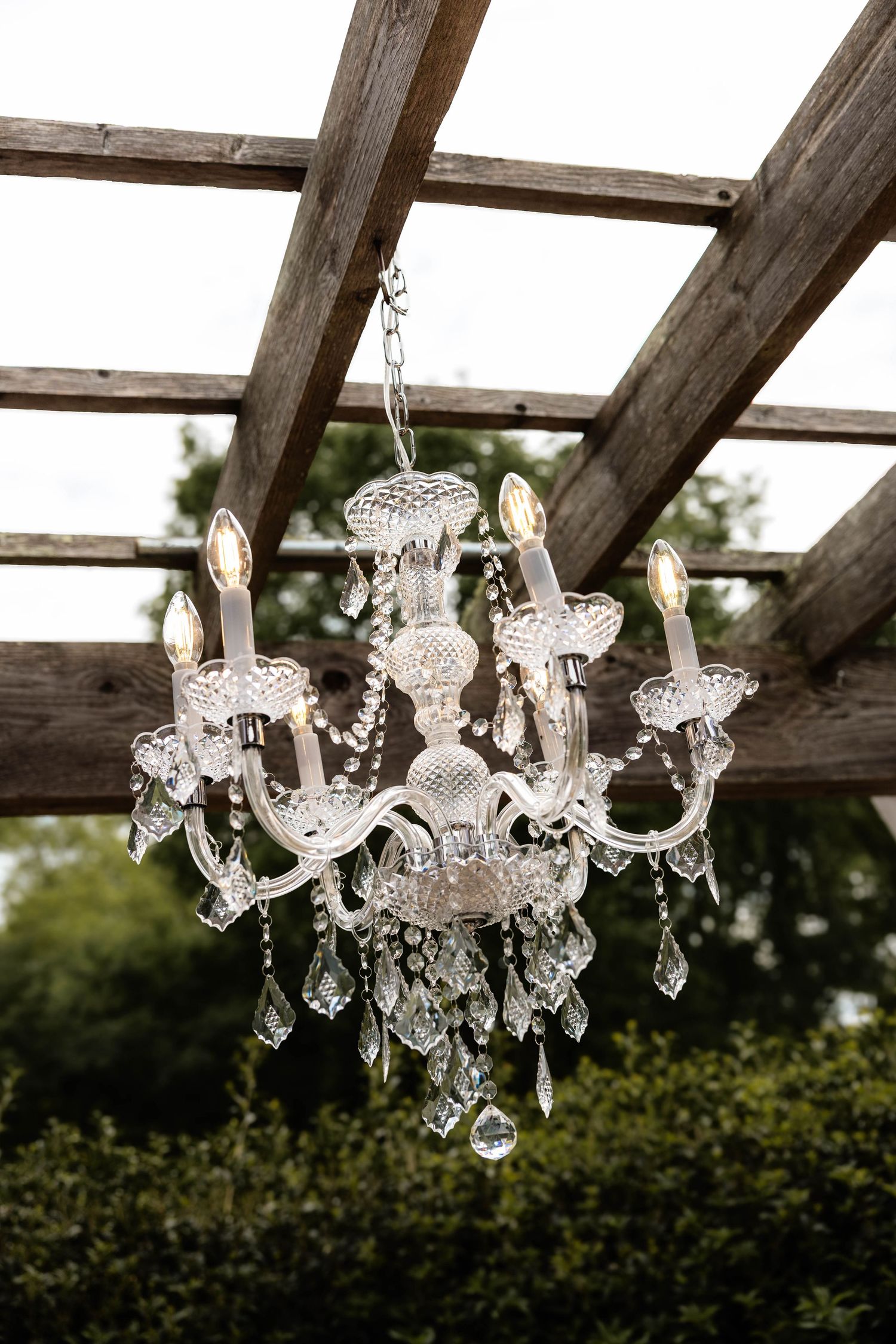 Elegant crystal chandelier hanging from a rustic wooden pergola outdoors, adorned with candle lights and sparkling glass pendants.