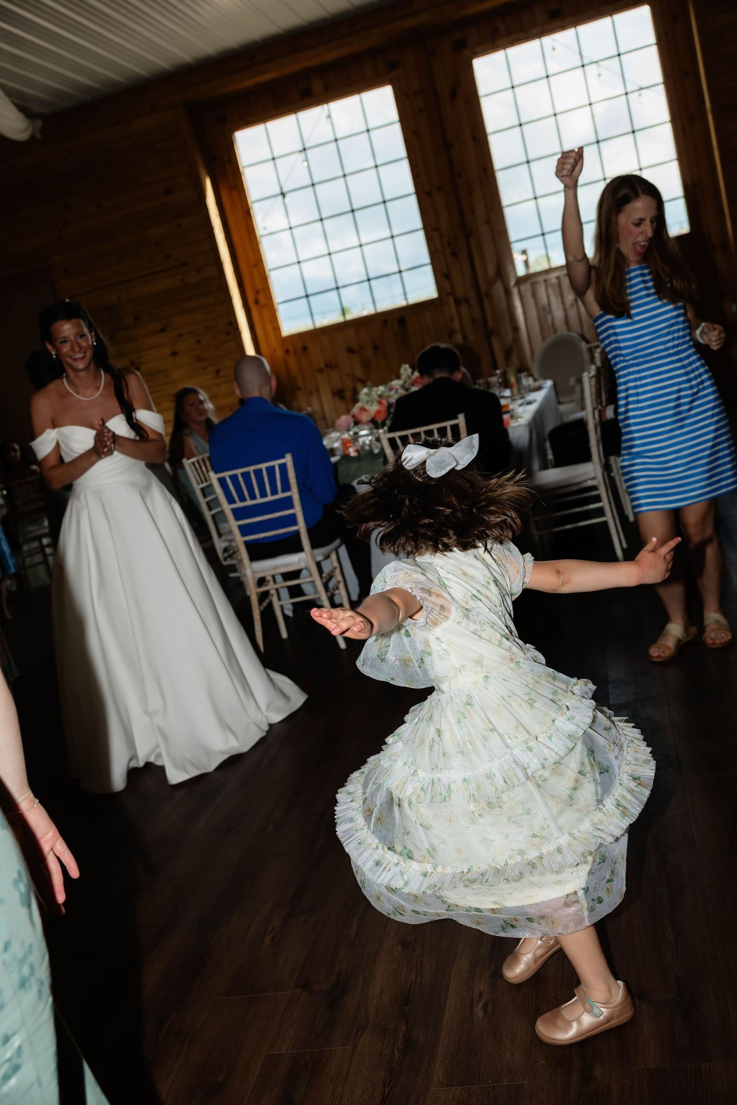 A young flower girl twirls in a floral dress on the dance floor at a rustic wedding reception venue.
