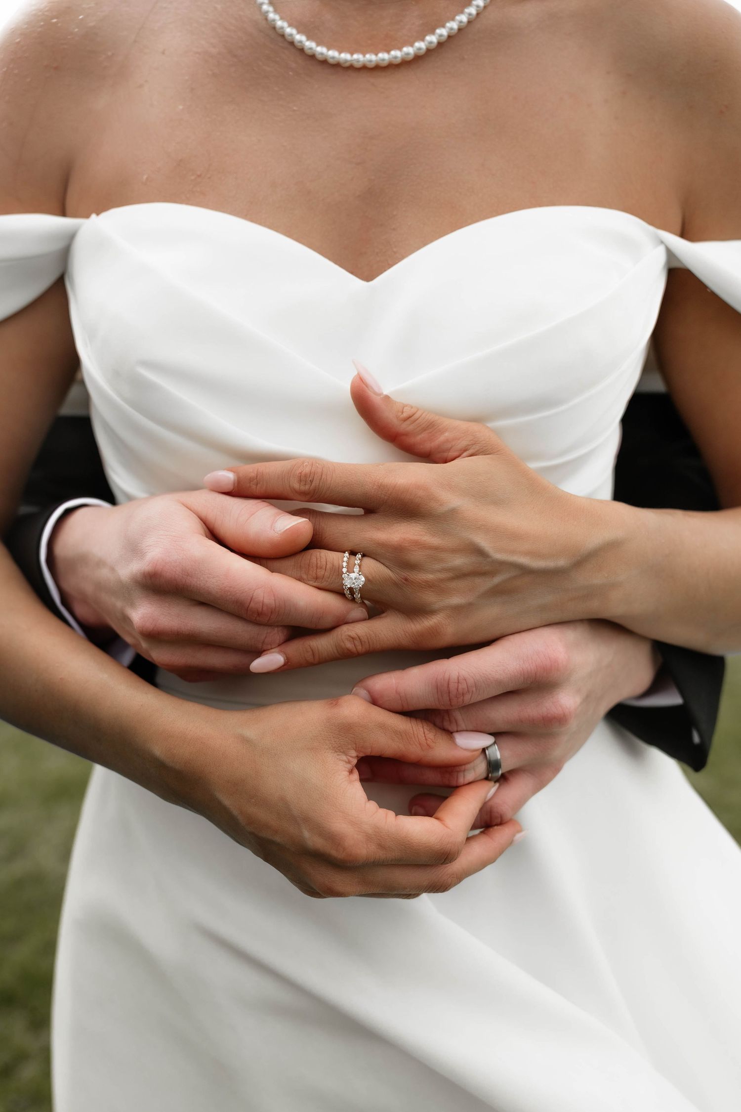 Bride and groom holding hands showing wedding rings, bride wearing white off-shoulder gown and pearl necklace.