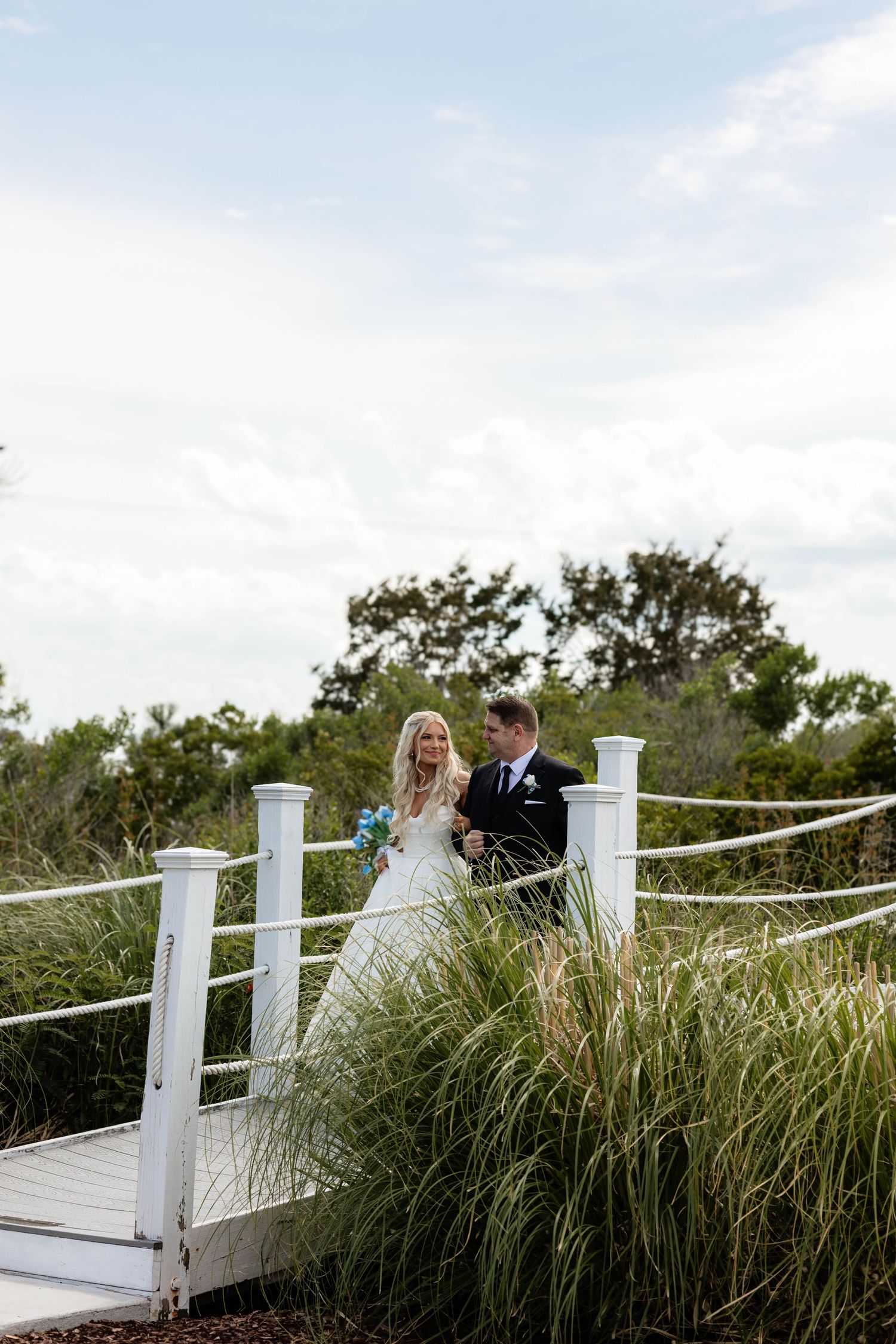 Bride and groom walking on a white wooden bridge surrounded by tall grass on their wedding day.