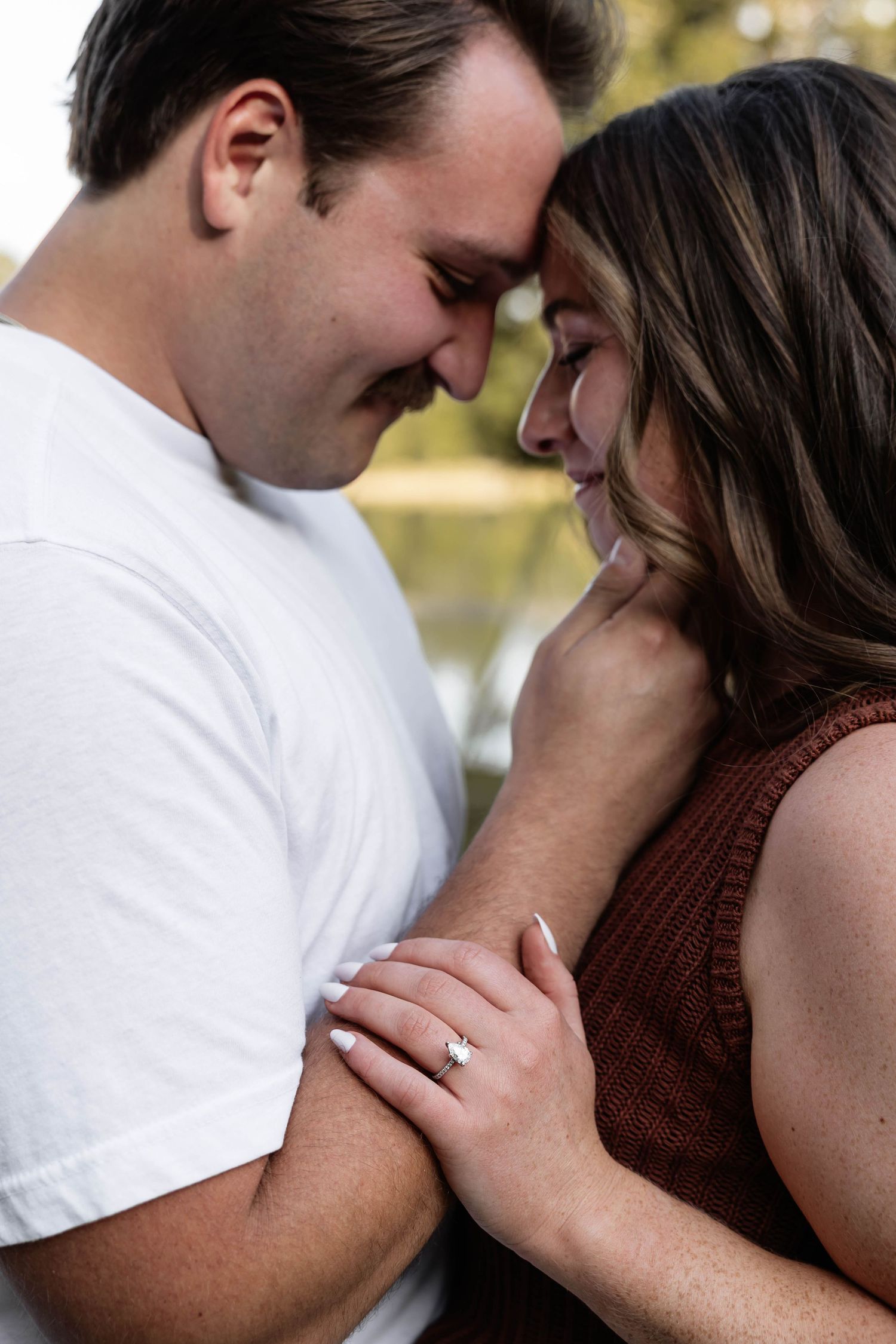 Romantic couple touching foreheads and smiling, woman displaying engagement ring, outdoor bokeh background.