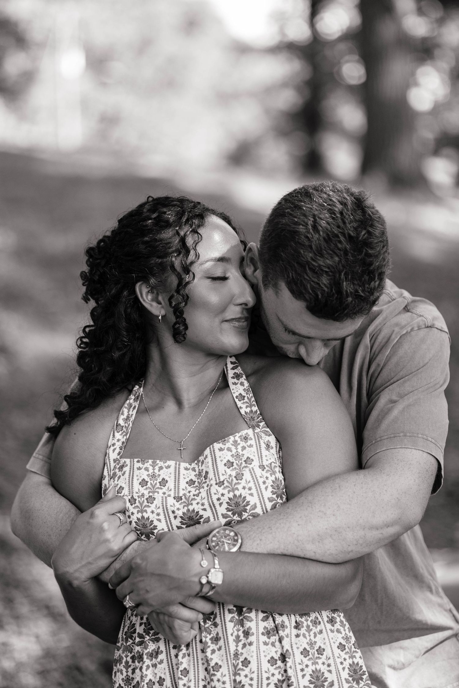 Black and white photo of a couple embracing outdoors, man nuzzling woman's cheek as she smiles softly.