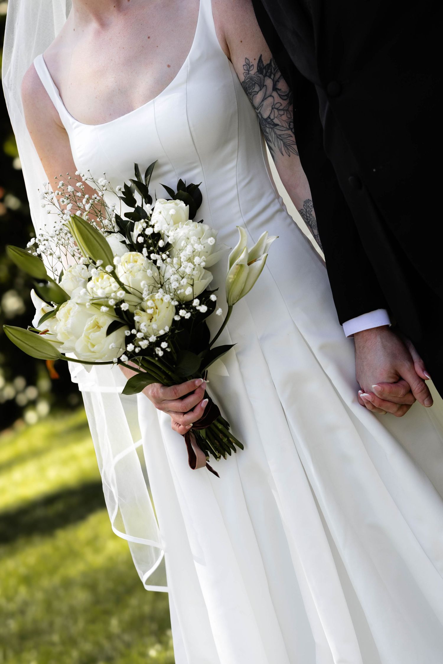 Bride in white gown holding white floral bouquet with lilies, standing beside groom in black suit outdoors.