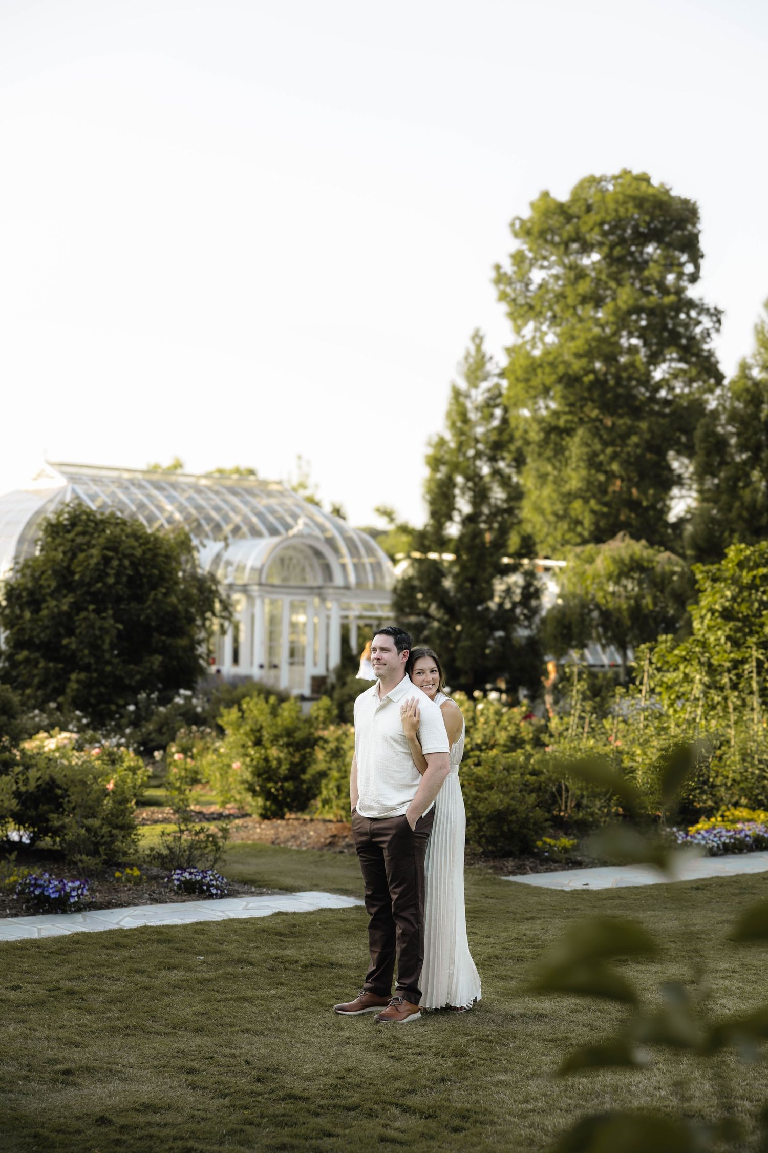Couple in formal attire embracing in lush garden with greenhouse backdrop during golden hour.