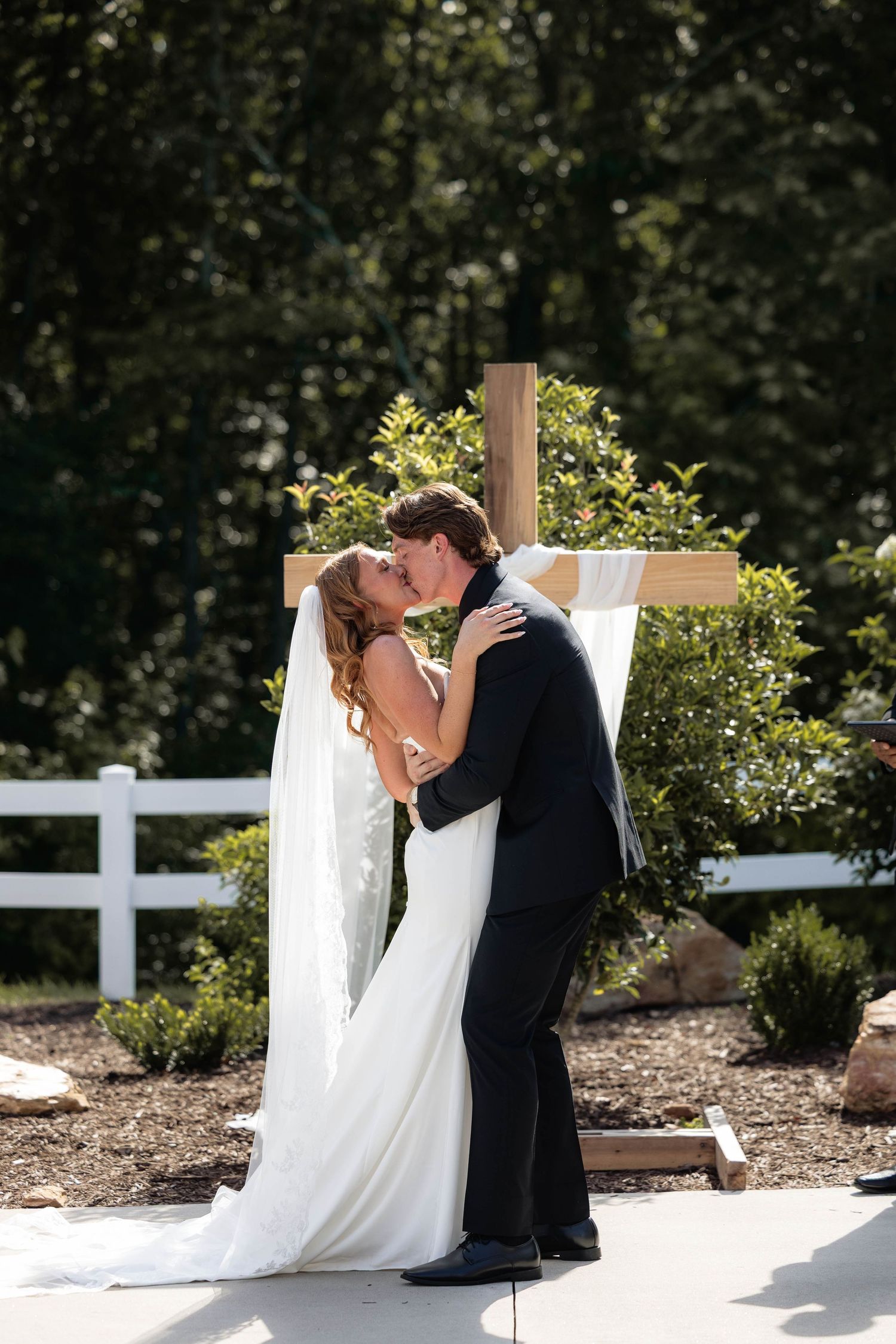 Bride and groom share first kiss at outdoor wedding ceremony in front of wooden cross surrounded by lush greenery.