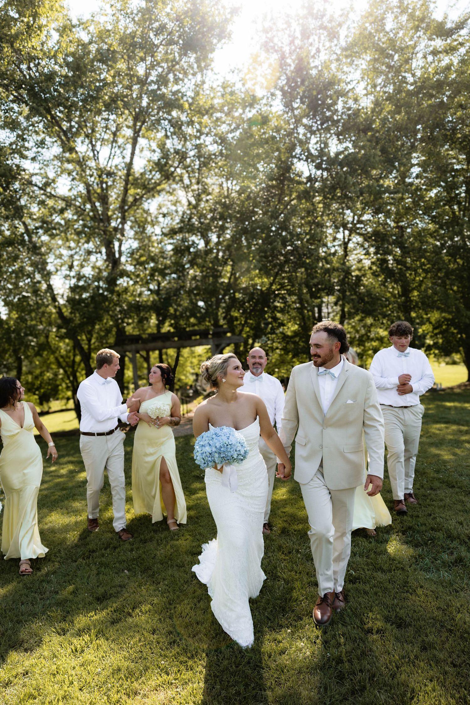 Bride and groom hold hands walking with wedding party in yellow dresses and white suits on sunny green lawn.
