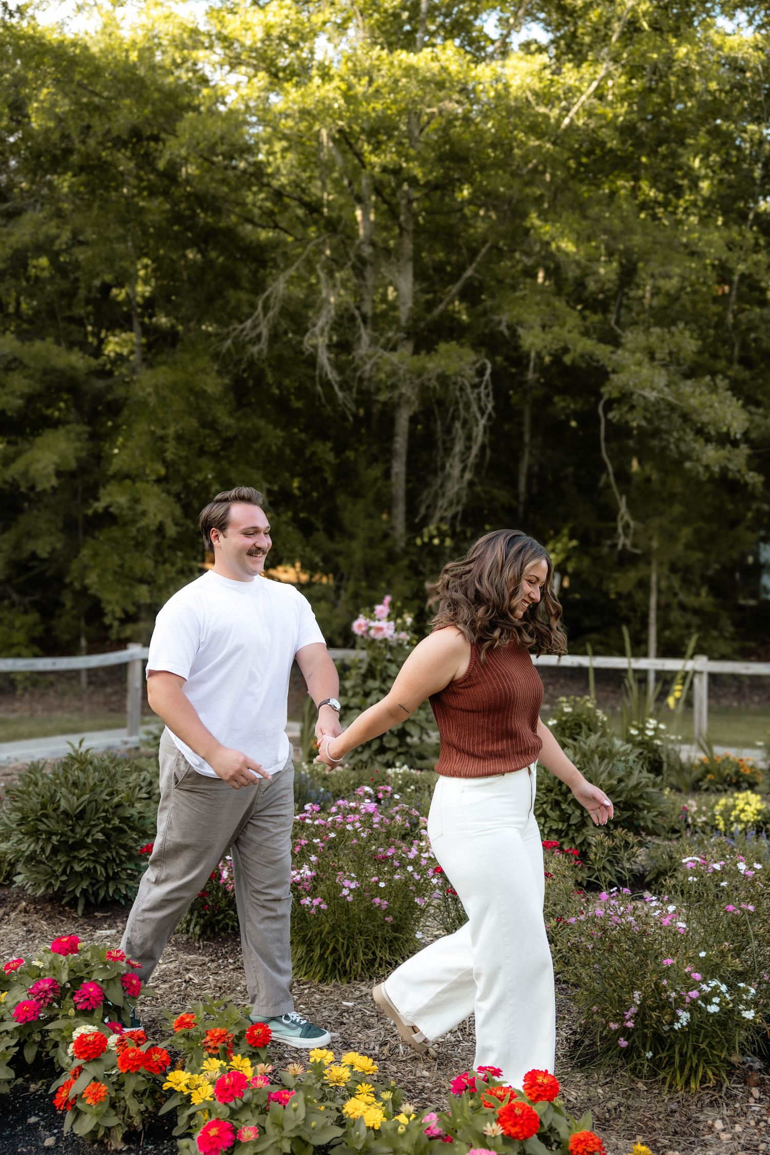 Couple holding hands and walking through a colorful flower garden with lush green trees in the background.