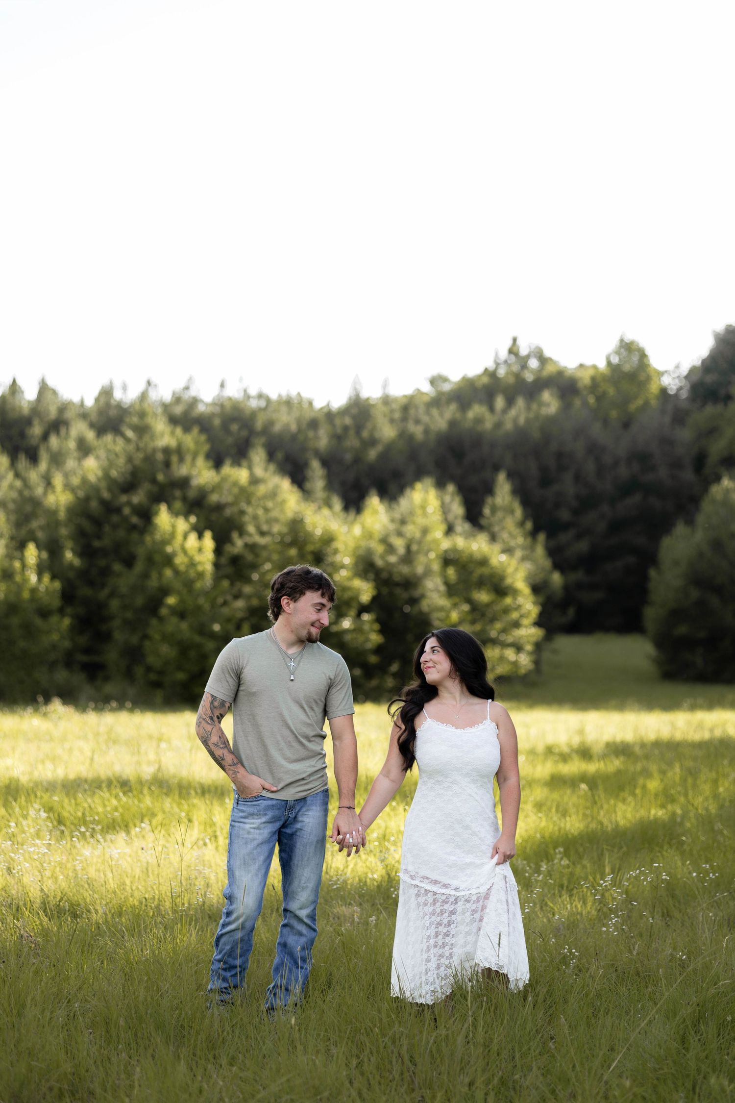 A couple holds hands in a green meadow, smiling at each other with a forest backdrop on a sunny day.