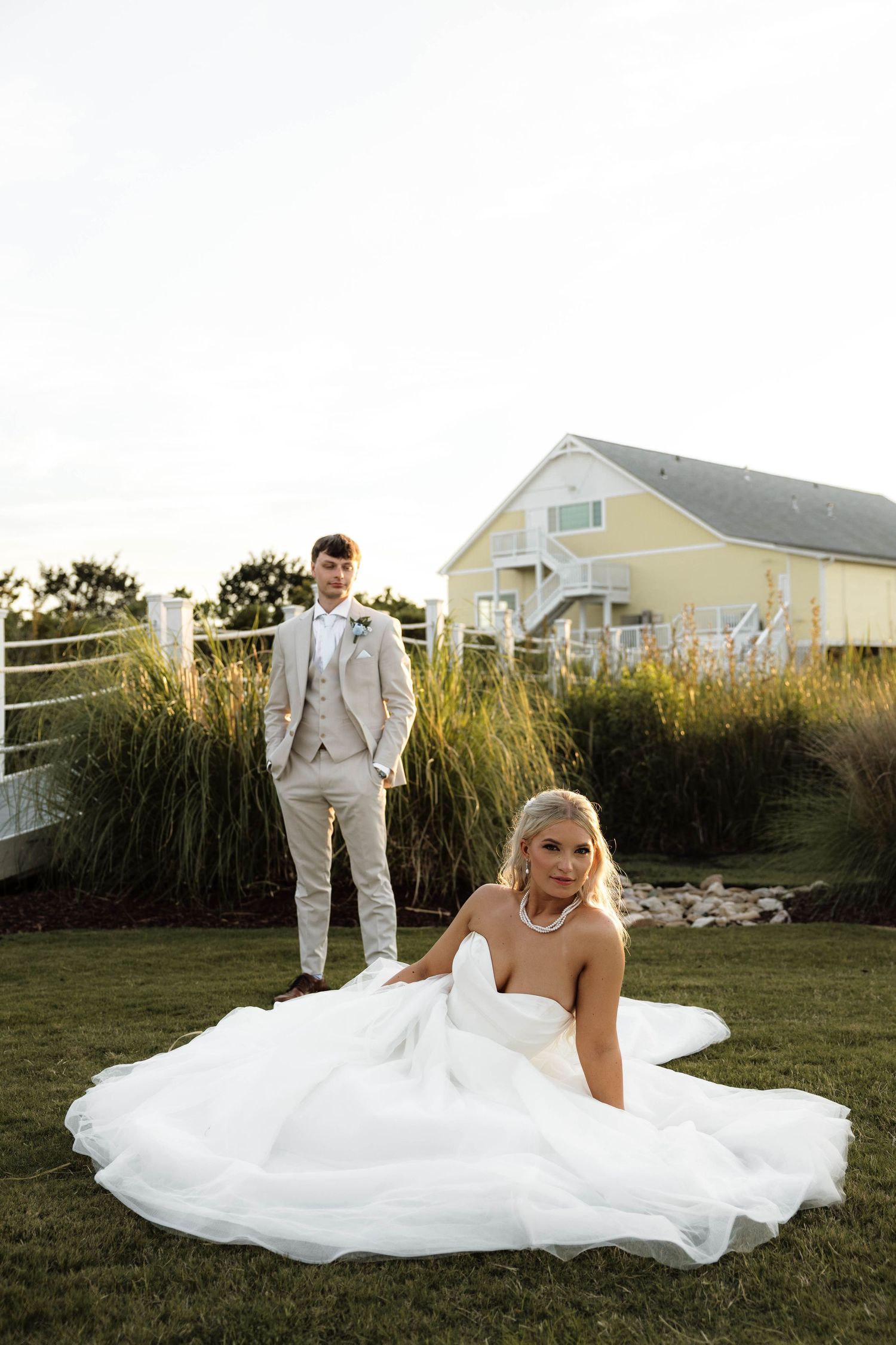 Bride in a flowing white gown sits on grass while groom stands behind her near a yellow coastal home.