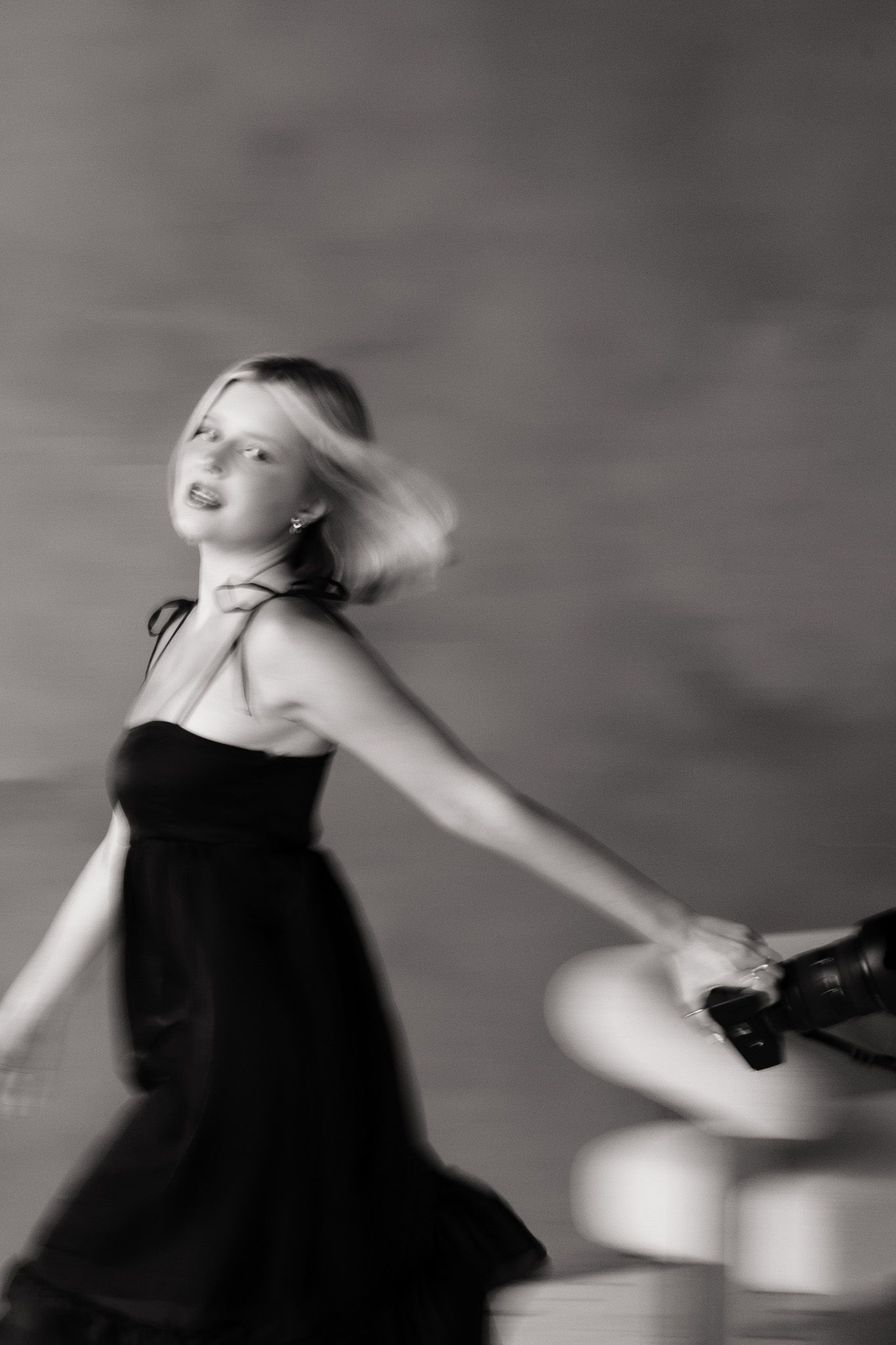 Black and white photo of a blonde woman in a black dress holding a camera, captured with motion blur effect.