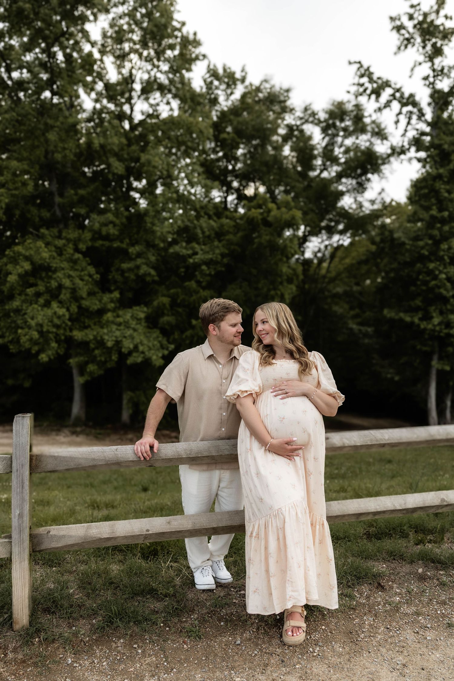 Pregnant couple posing by a wooden fence surrounded by lush green trees, woman in a flowing peach dress cradling her baby bump.