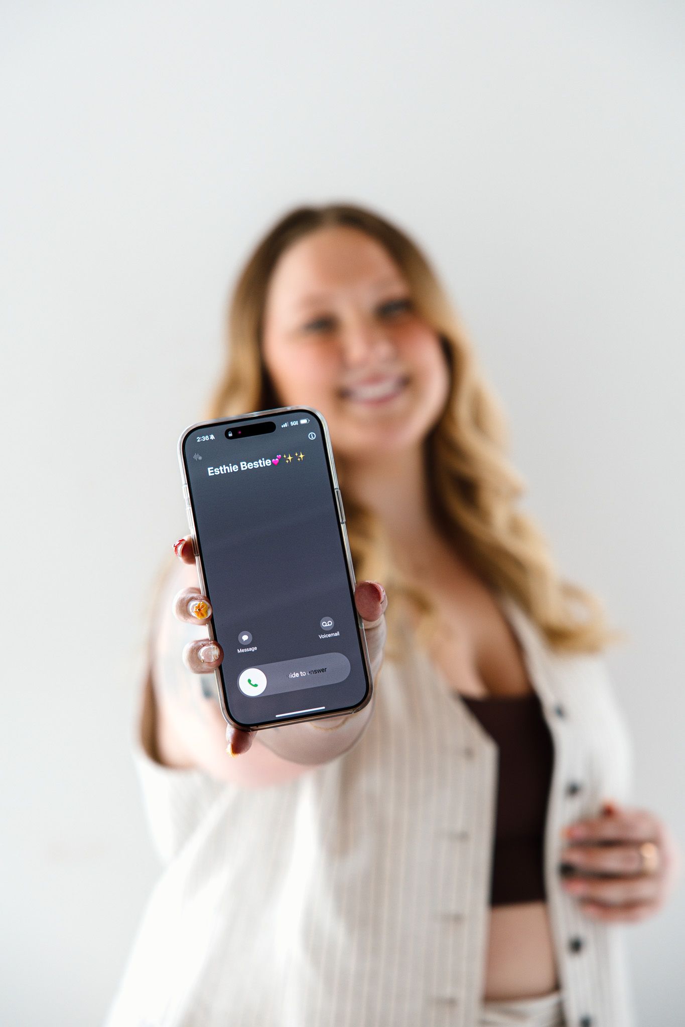 Smiling blonde woman holding iPhone displaying a phone call screen toward camera against white background.