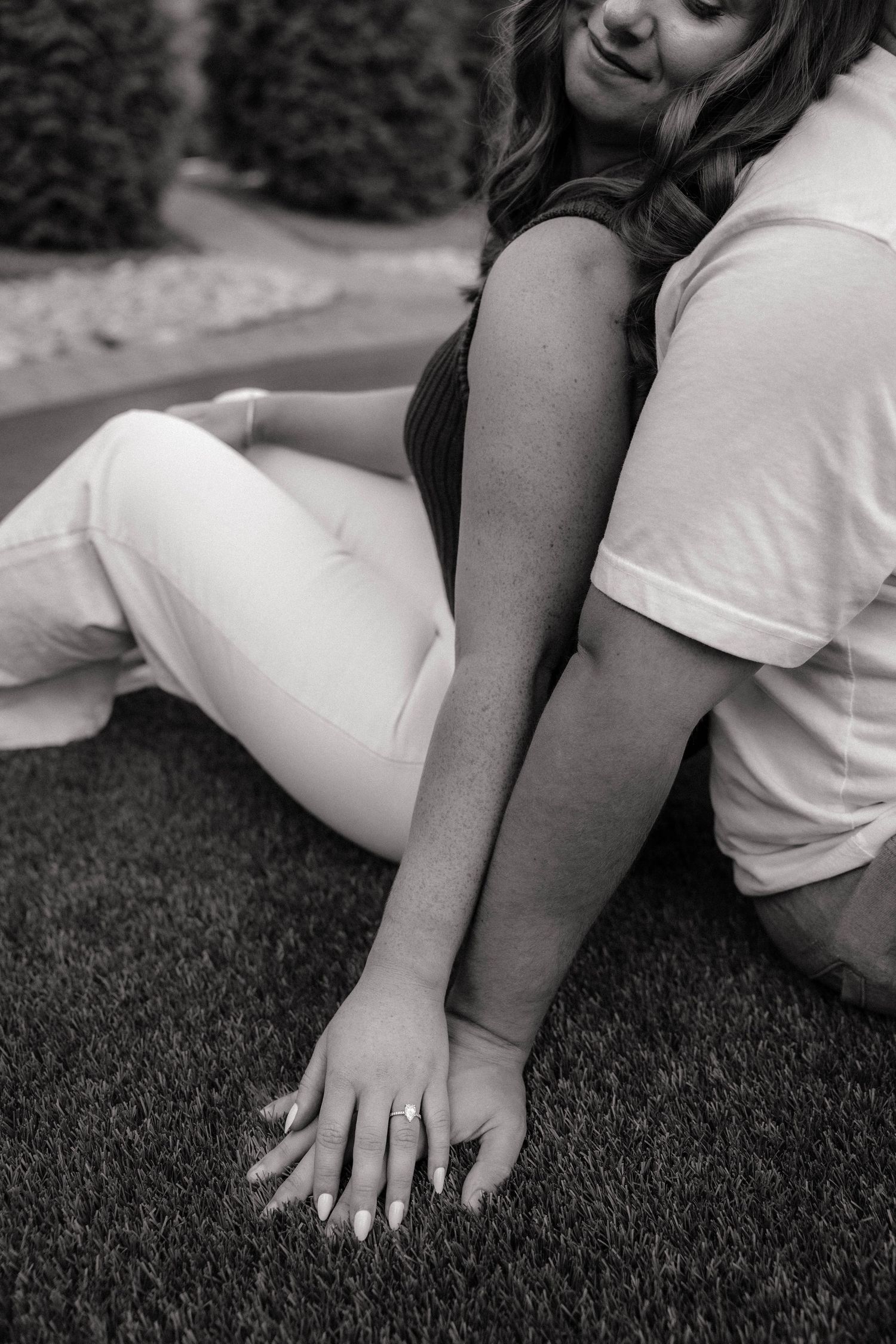 Black and white photo of couple sitting back to back on grass, woman showing engagement ring on her hand.