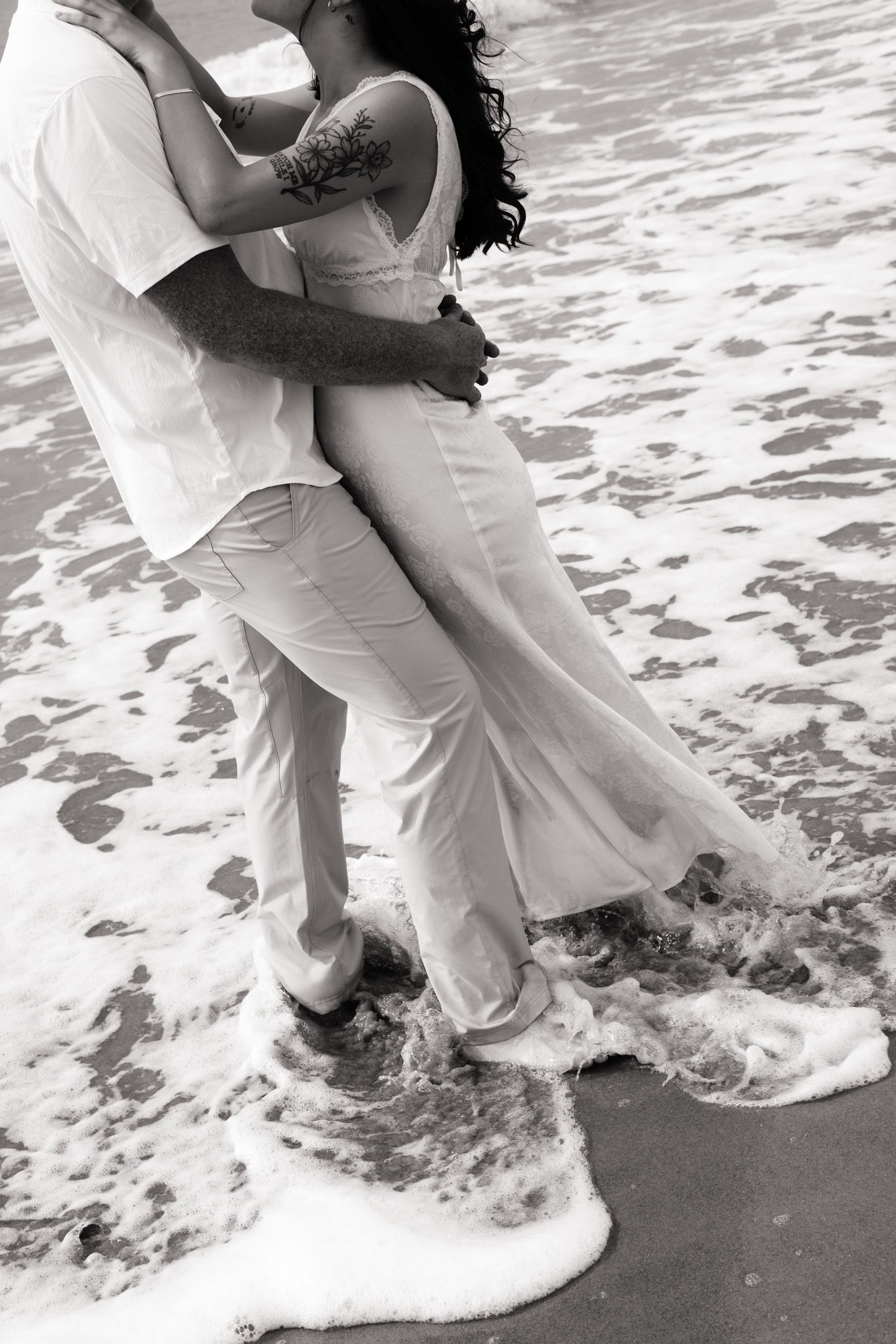 Black and white photo of a couple embracing on a beach, waves washing over their feet, woman in white dress.