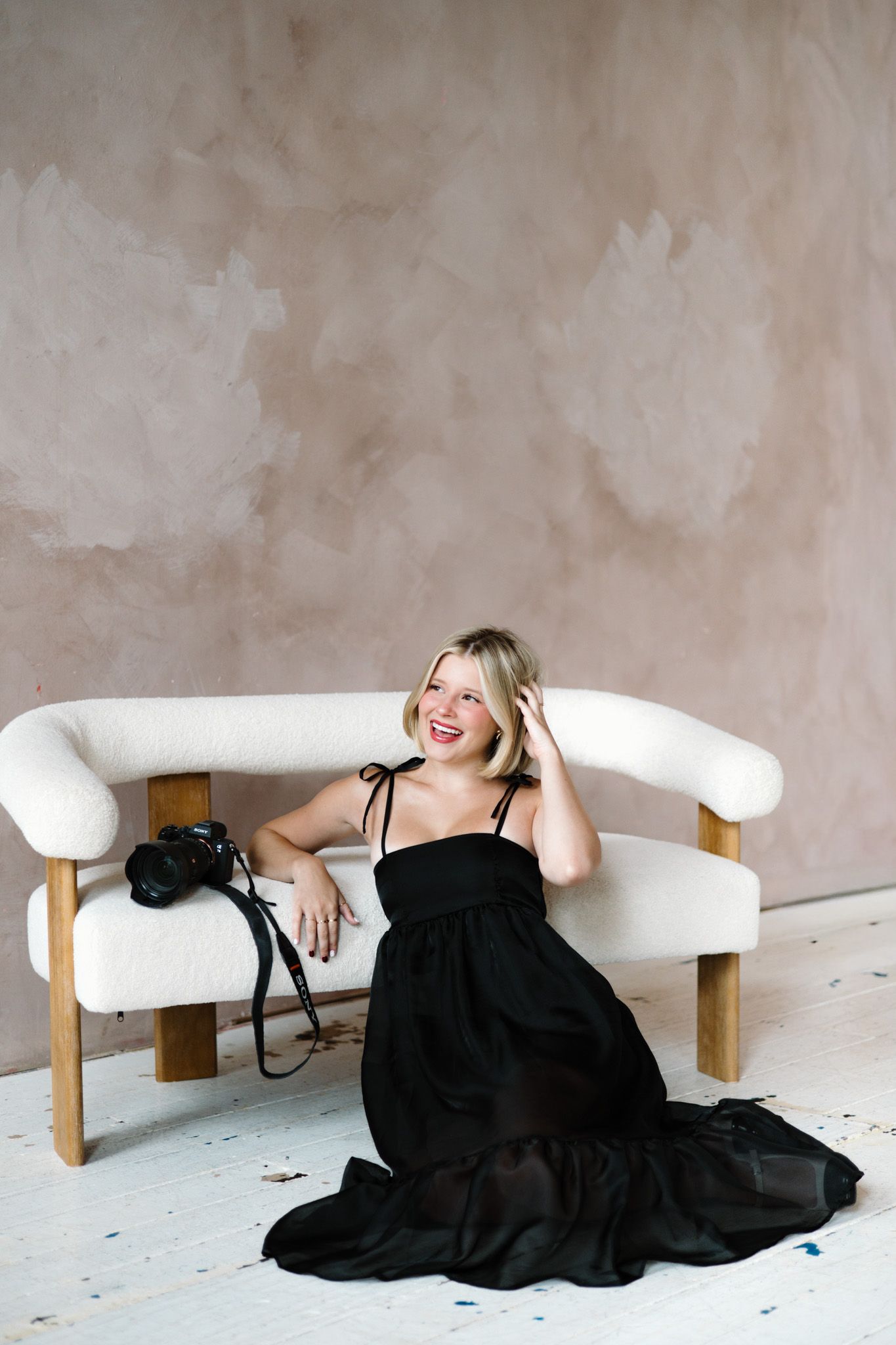Smiling blonde woman in black gown sits on white boucle sofa with camera, in a neutral studio setting.